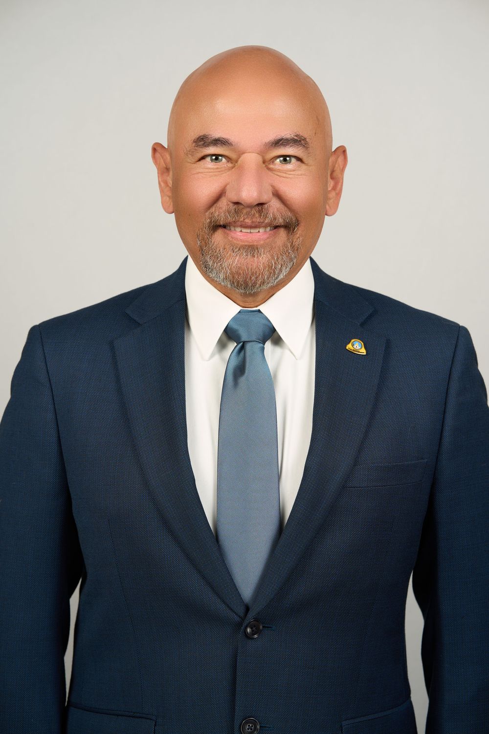 Professional headshot of a smiling bald man with beard wearing a navy suit and blue tie.