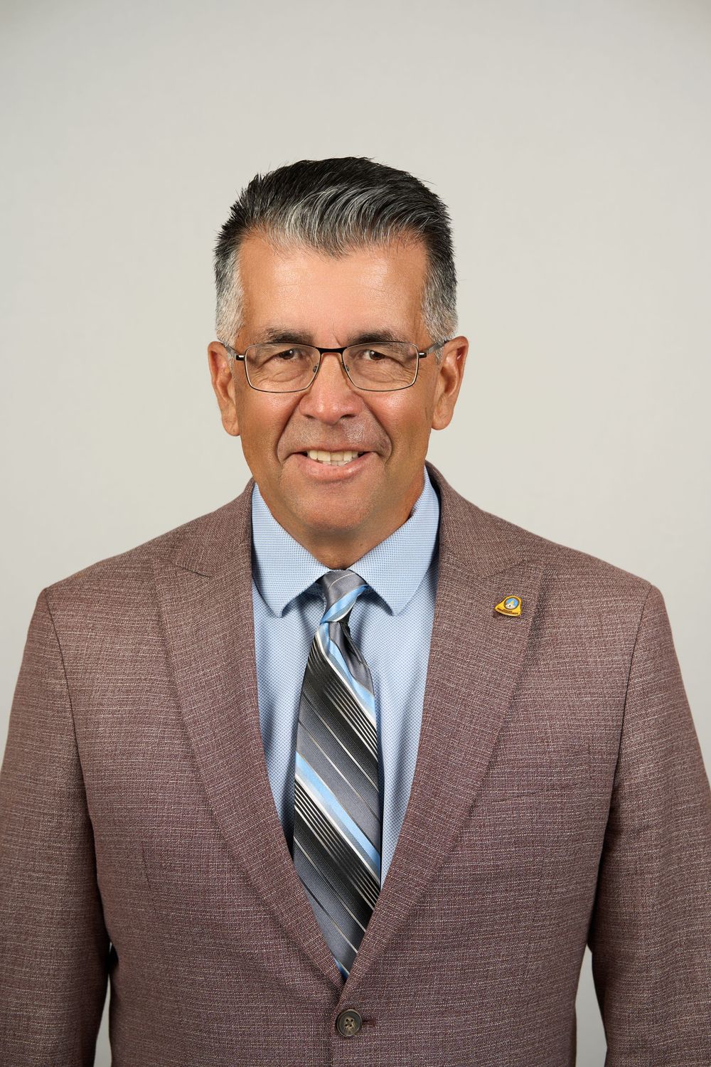 Professional headshot of a middle-aged man in glasses, wearing a brown suit and striped tie, smiling confidently.