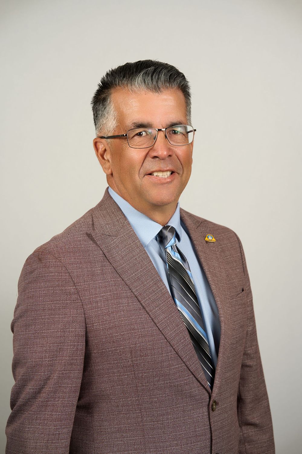 Professional headshot of a middle-aged man in glasses, brown blazer, and striped tie, smiling confidently.