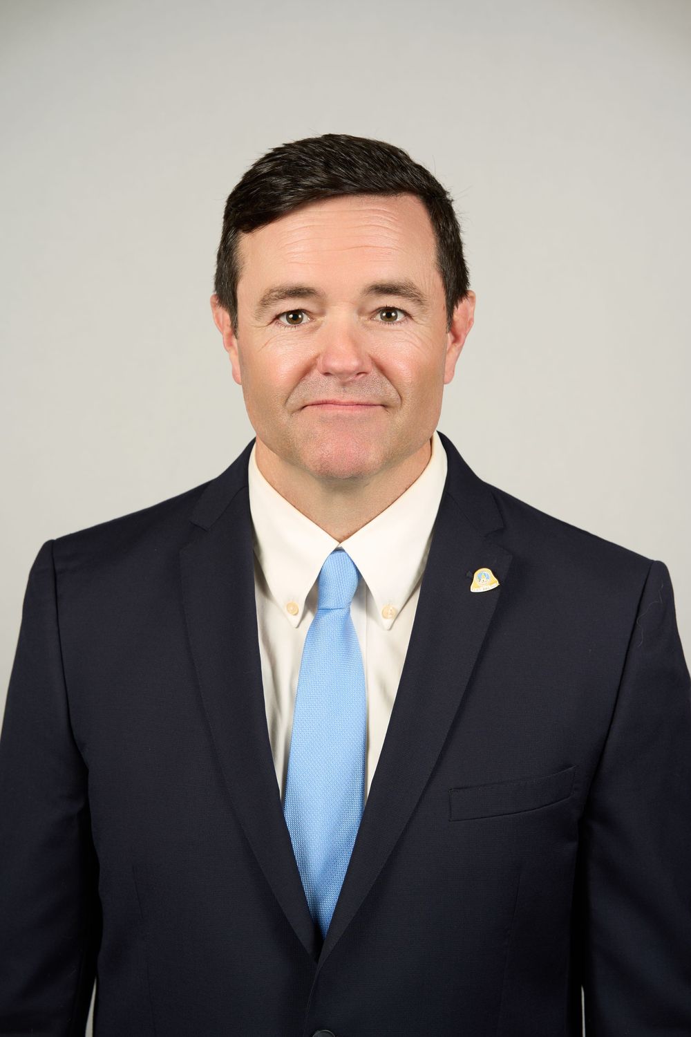 Professional headshot of a man in a navy suit and light blue tie smiling against a gray background.