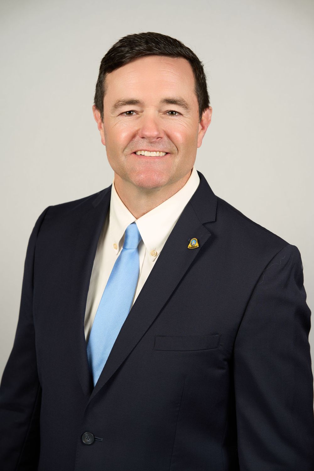 Professional headshot of a smiling man in a navy suit and light blue tie against a gray background.