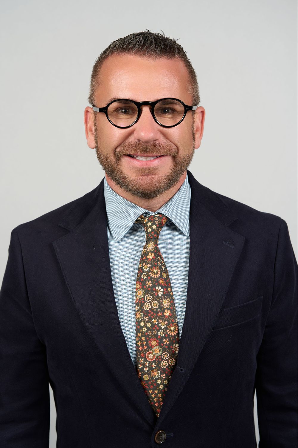 Professional man in dark suit, floral tie, and round glasses smiling for a corporate headshot photo.