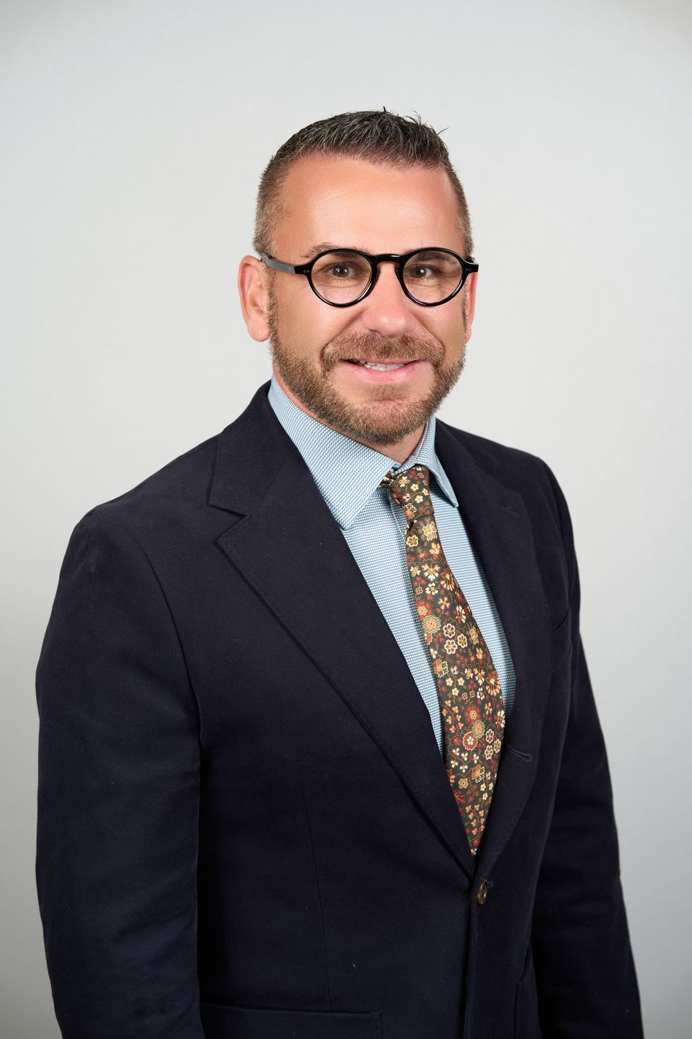 Professional man in dark suit, floral tie, and glasses smiling for a business headshot photo.