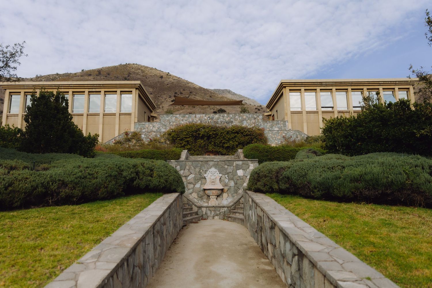 Elegant estate venue with manicured hedges, stone pathway, and mountain backdrop under cloudy sky.