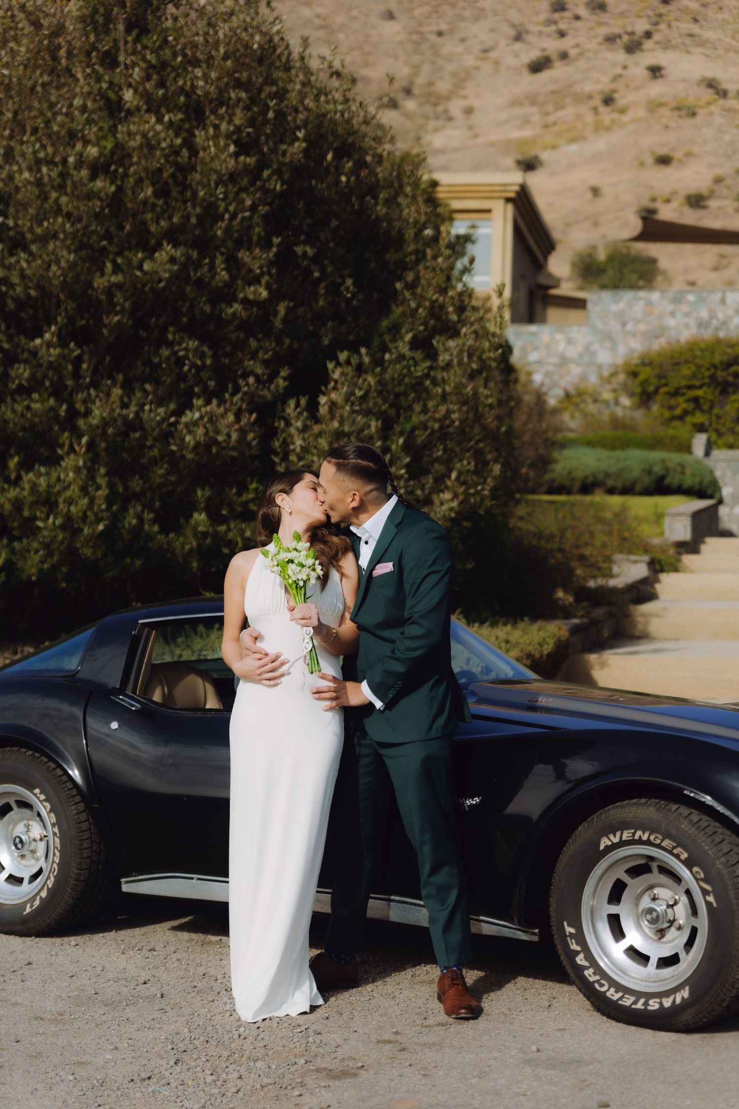 Bride and groom kissing beside a classic black Corvette with mountain views at their wedding.