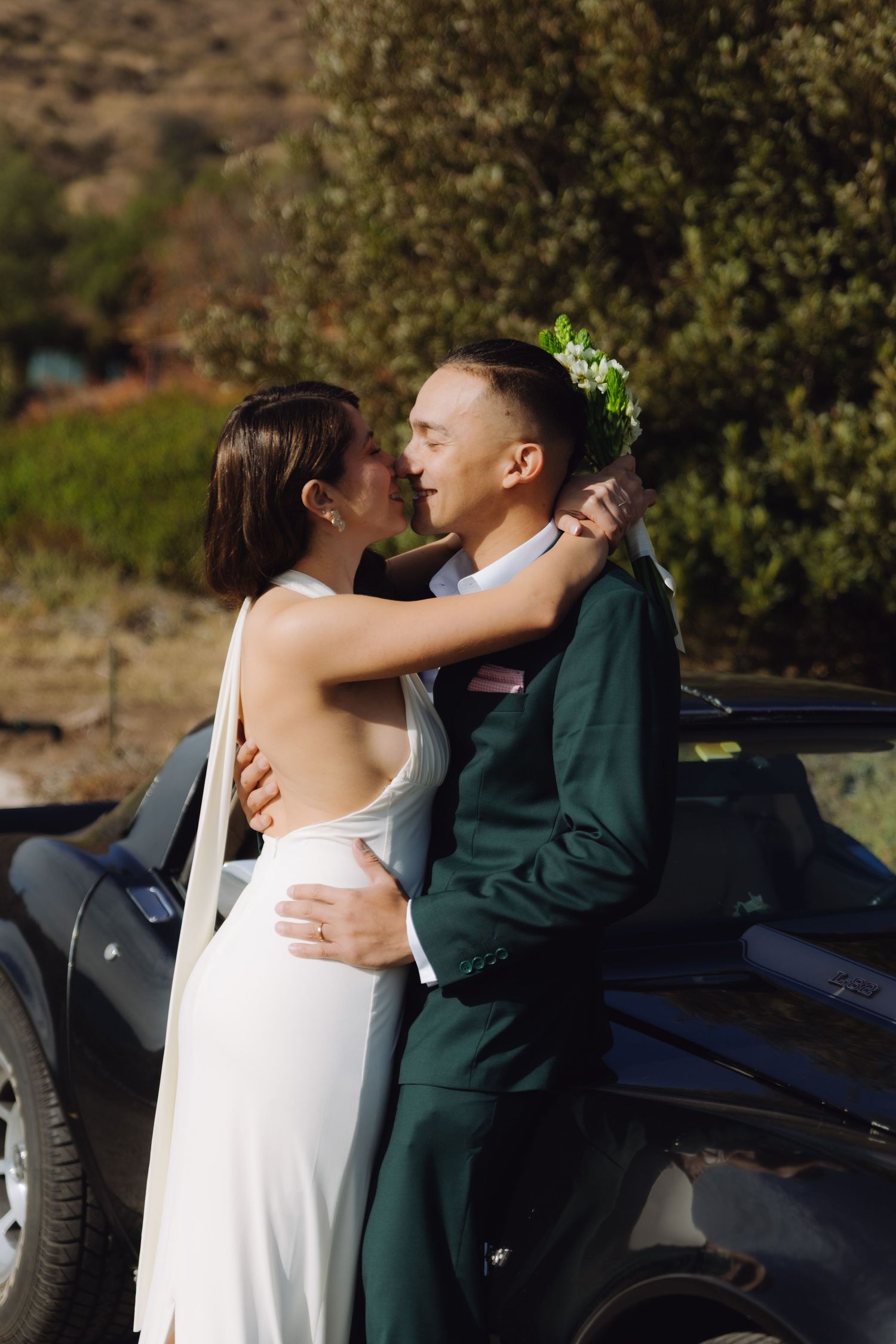 Wedding couple embracing and kissing beside a vintage black convertible in a scenic outdoor setting.