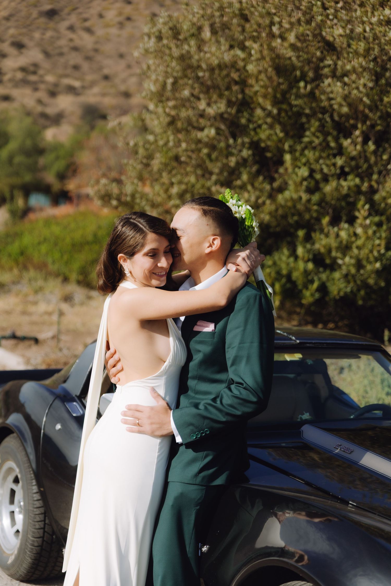 Newlyweds sharing a romantic embrace beside a vintage black car surrounded by lush green trees.