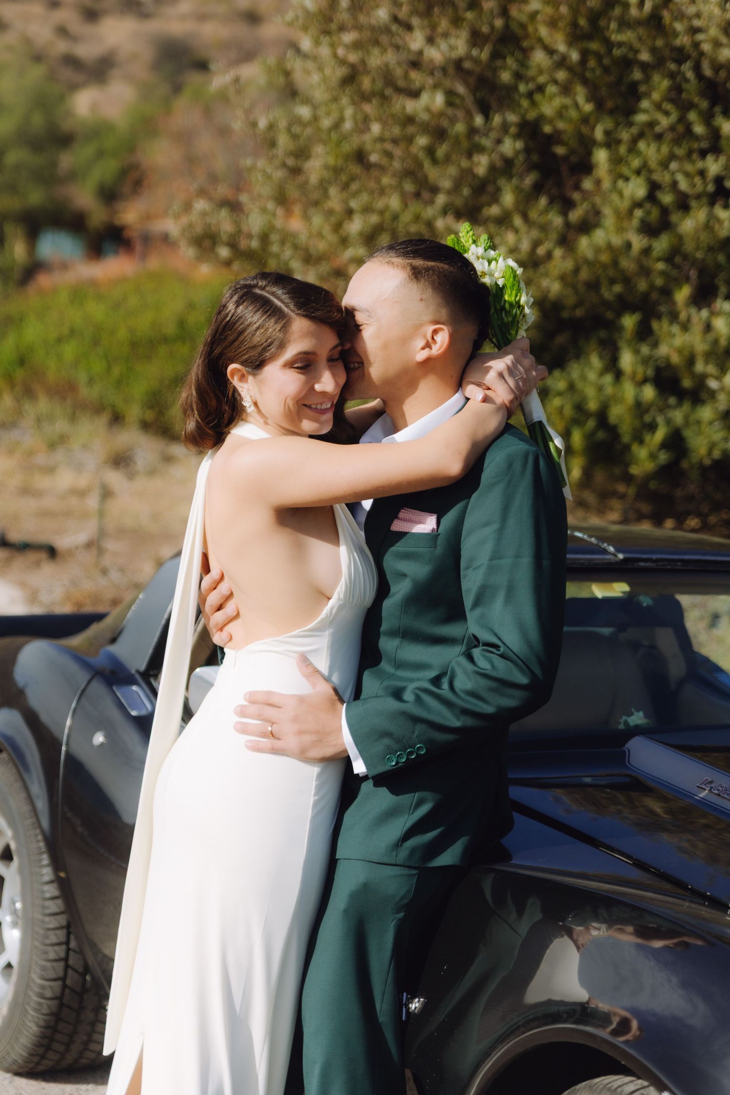 Smiling bride in white gown embracing groom in green suit beside a classic black car outdoors.