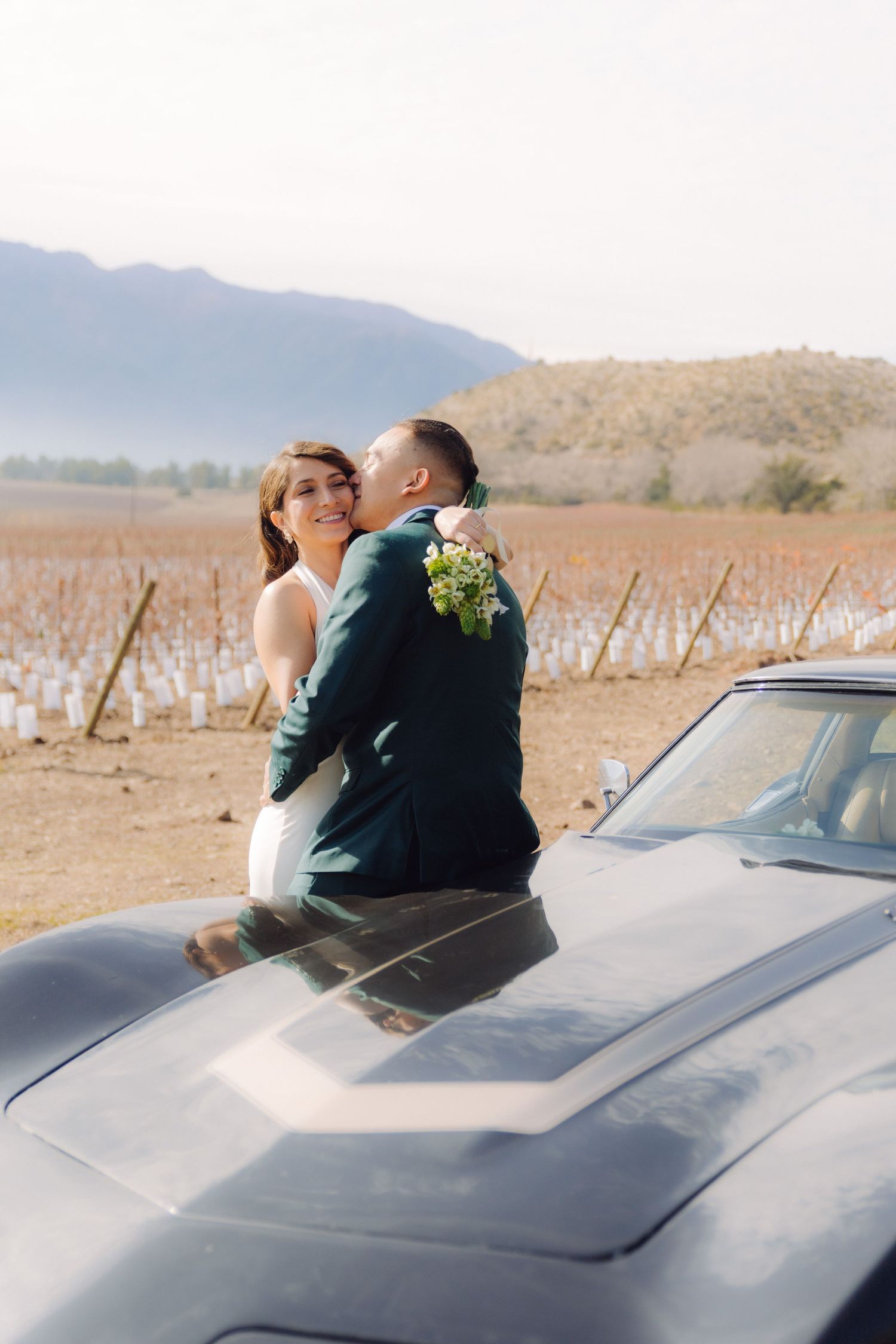 Bride and groom sitting on a classic black car hood embracing with vineyard and mountains behind.