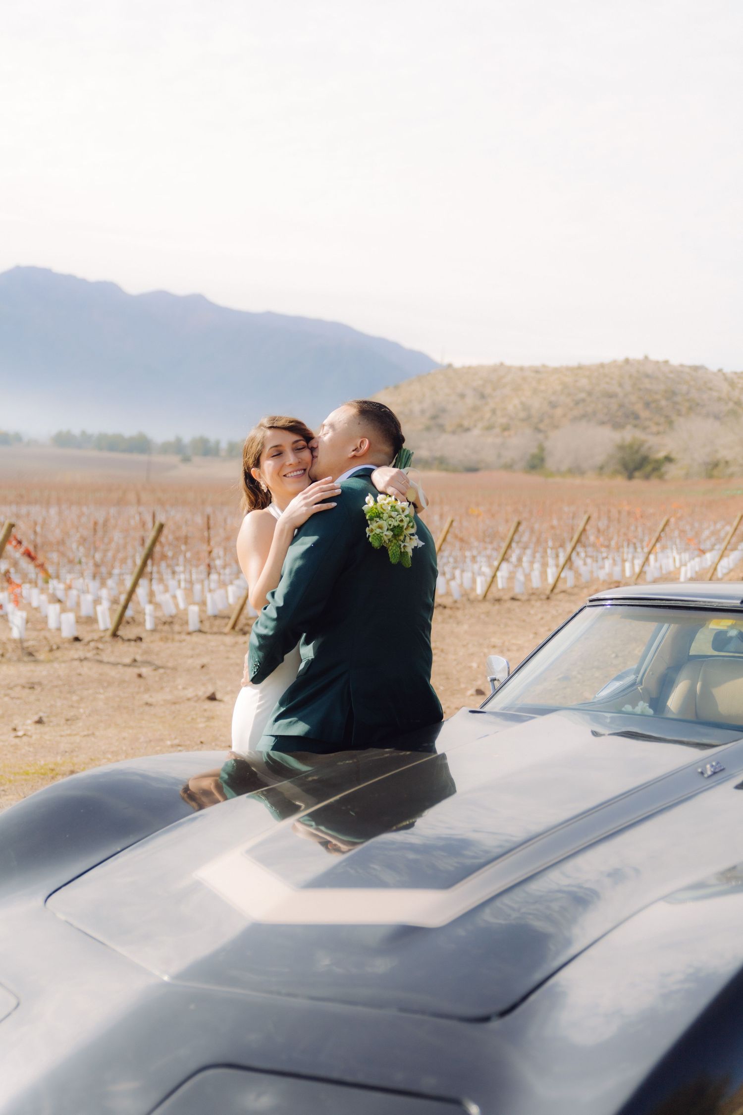 Wedding couple kissing on hood of vintage black car with scenic vineyard and mountain landscape.
