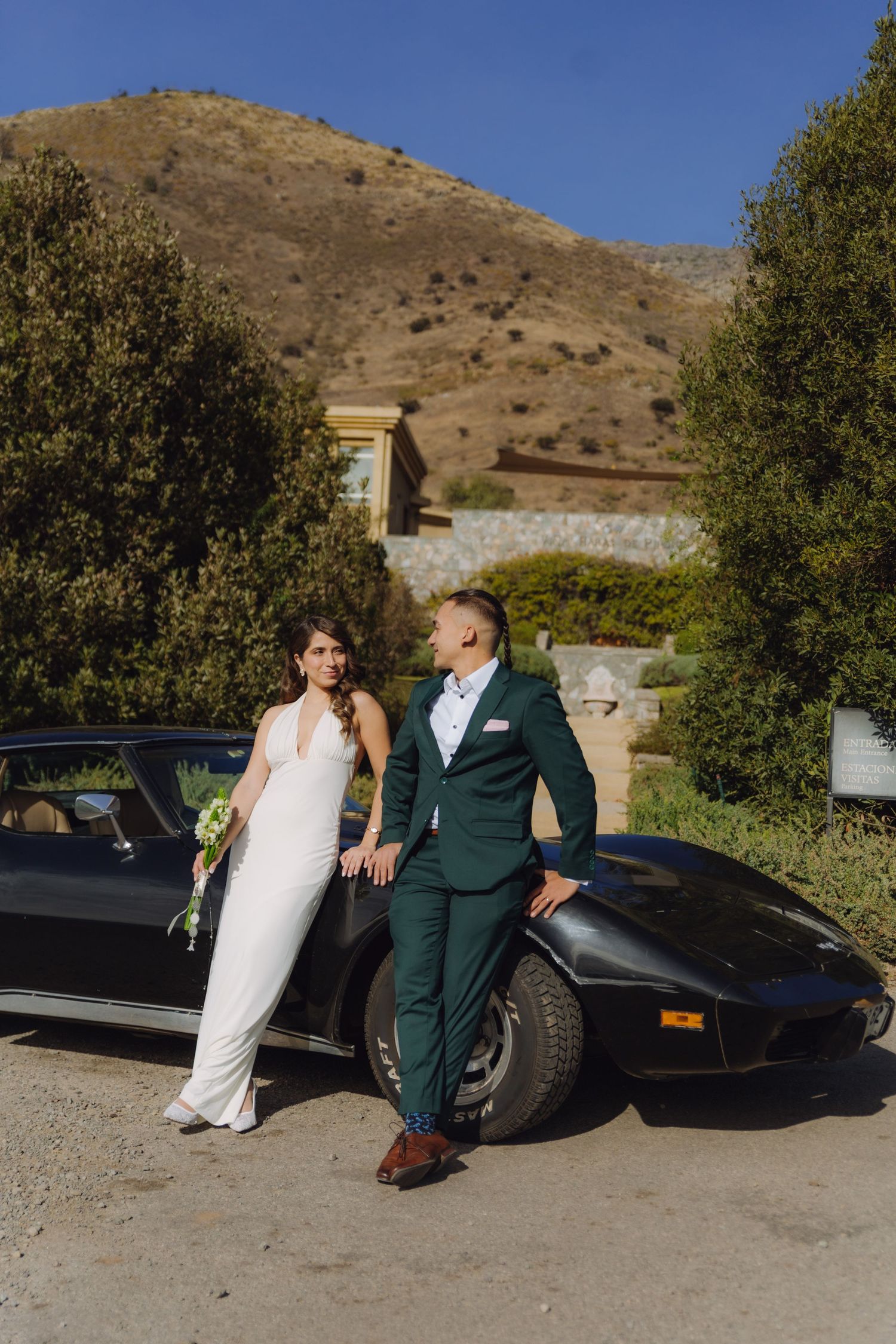 Bride in white halter gown and groom in green suit posing beside black Corvette at estate venue.