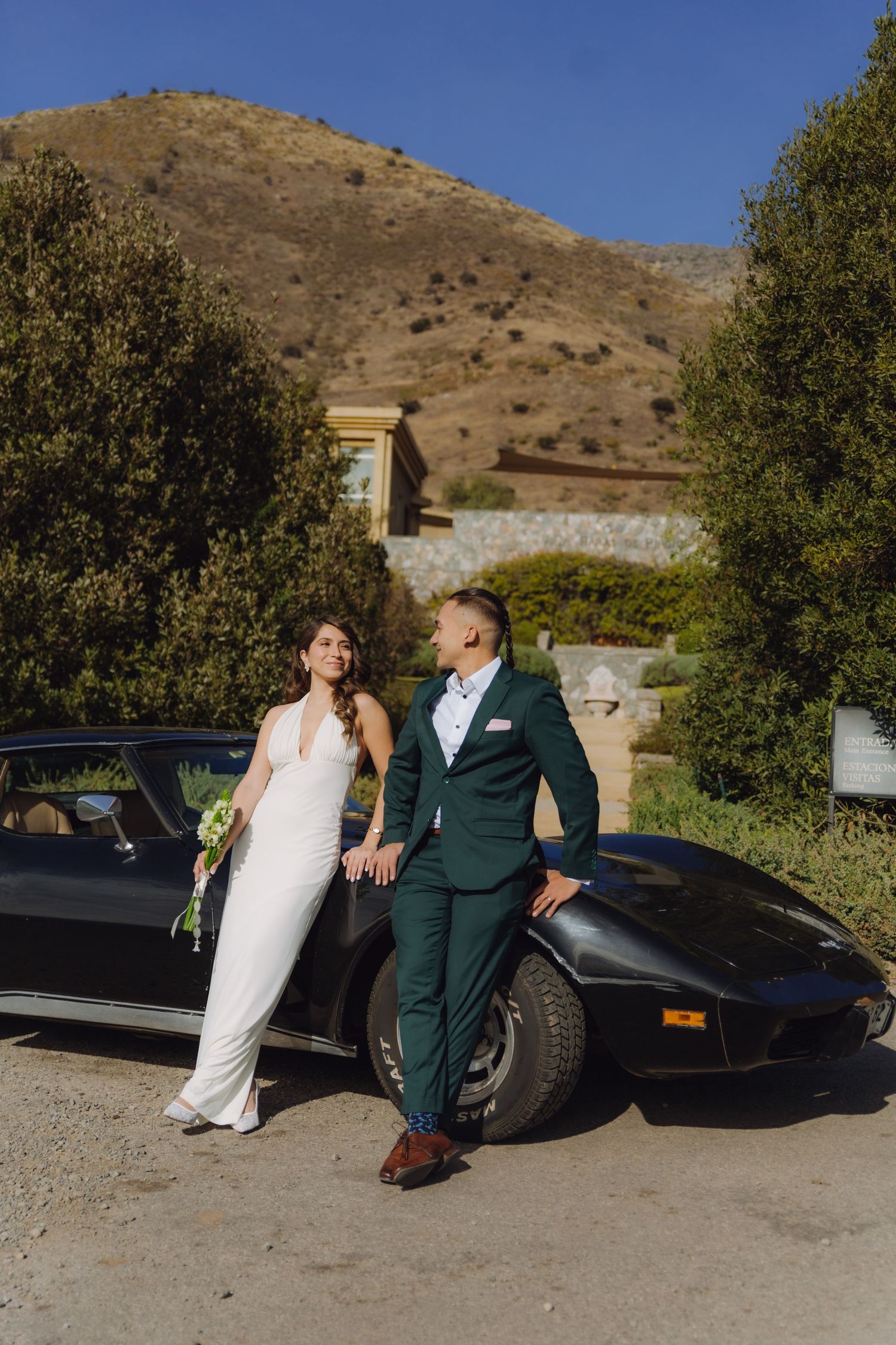 Newlyweds holding hands and smiling beside a classic black Corvette with hillside estate backdrop.
