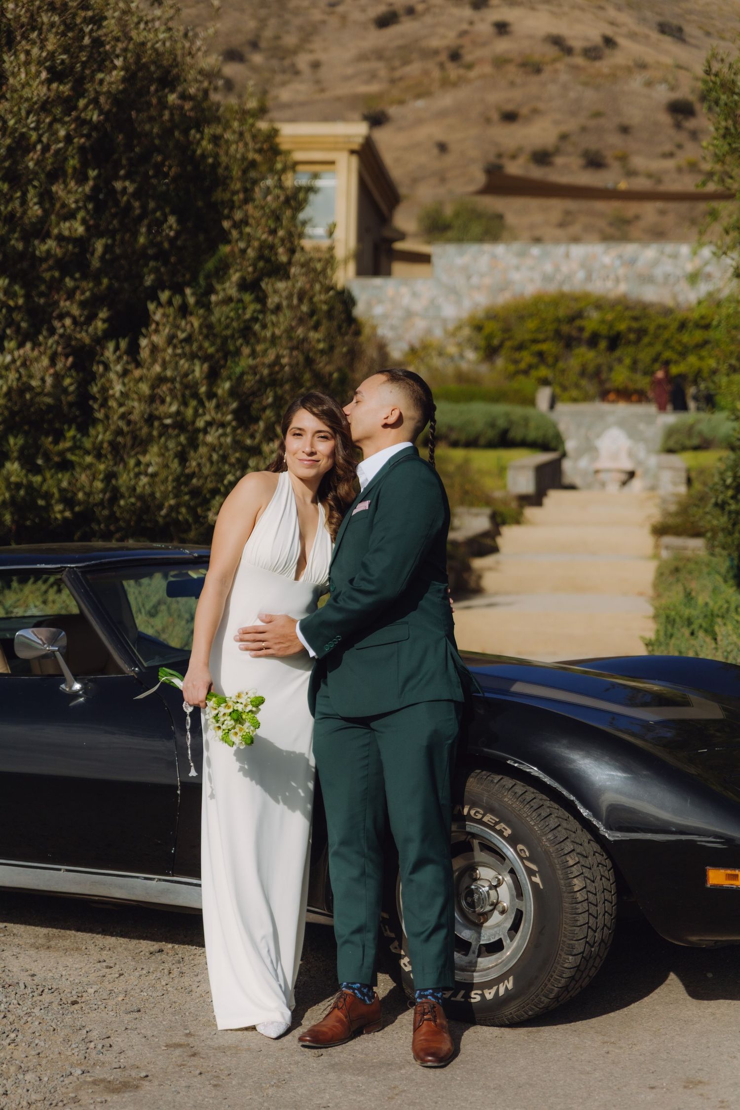 Groom in green suit kissing bride on cheek beside vintage black Corvette at elegant estate venue.