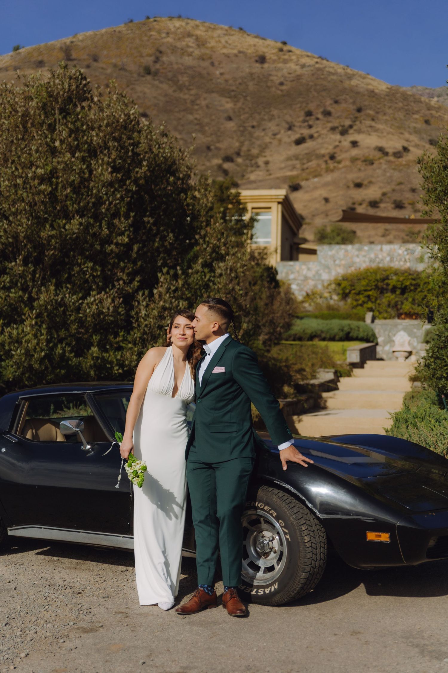 Bride and groom kissing beside a black vintage Corvette with a scenic mountain backdrop.