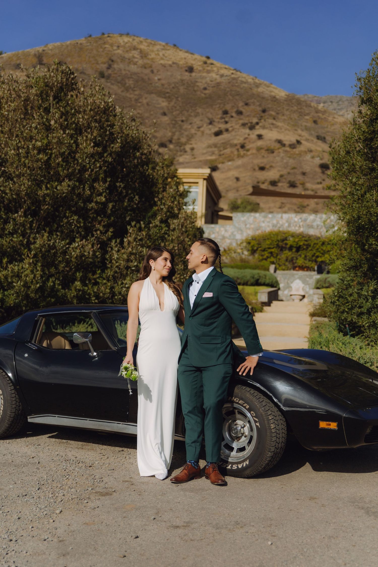 Wedding couple sharing a romantic gaze beside a classic black Corvette with mountain views.