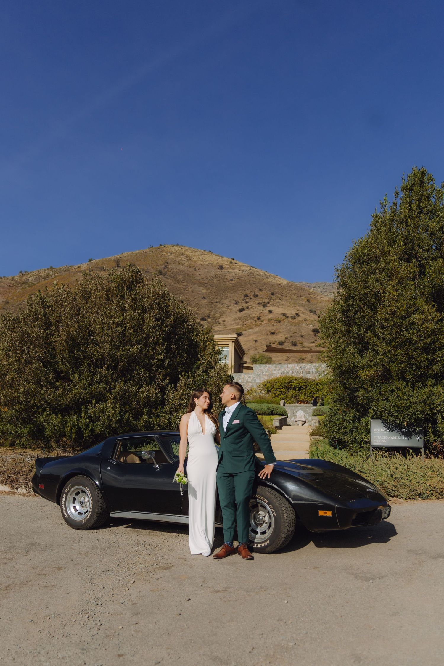 Bride and groom posing together beside a black vintage Corvette under a deep blue sky.