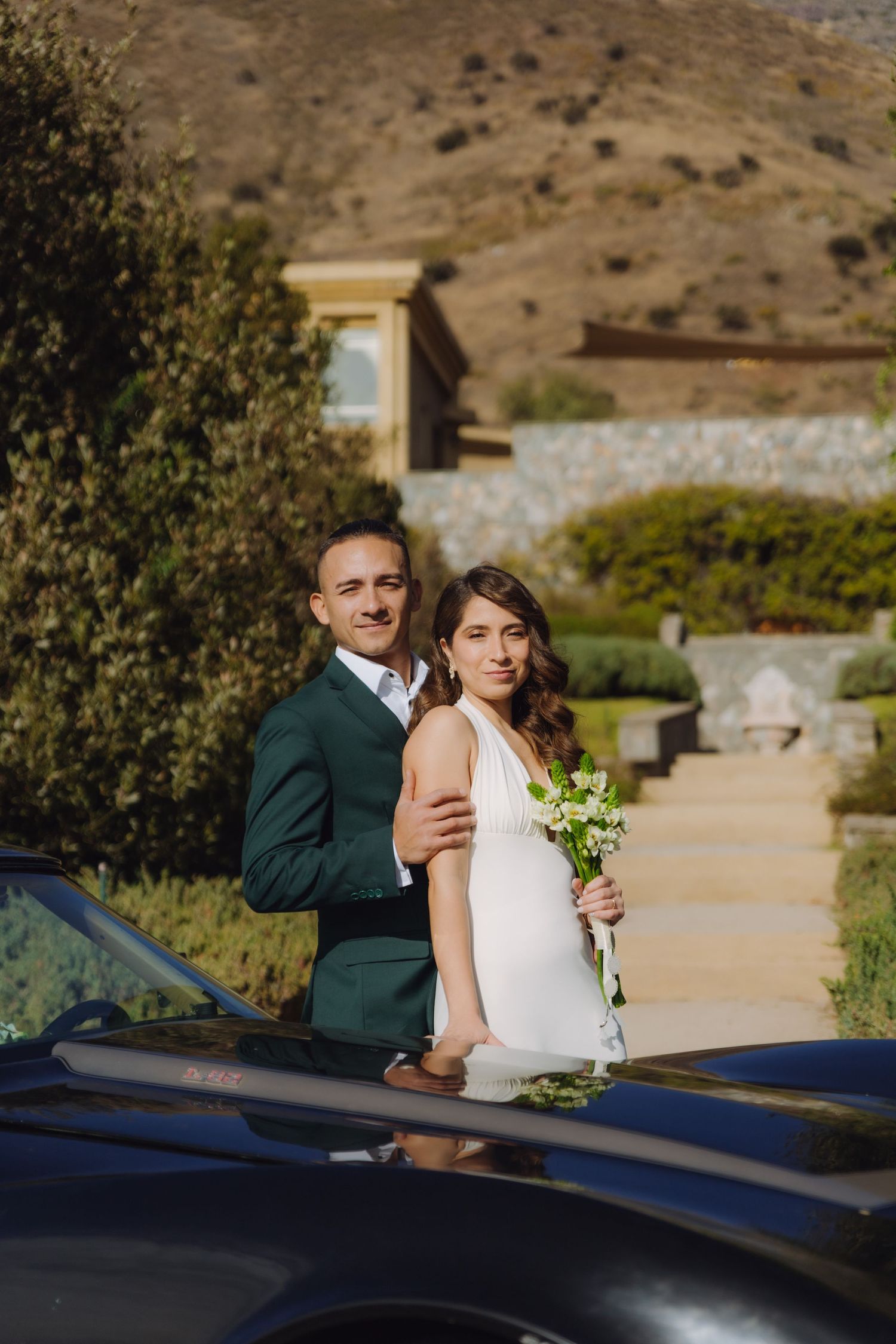 Bride and groom posing together beside a black classic car with a hillside estate backdrop.