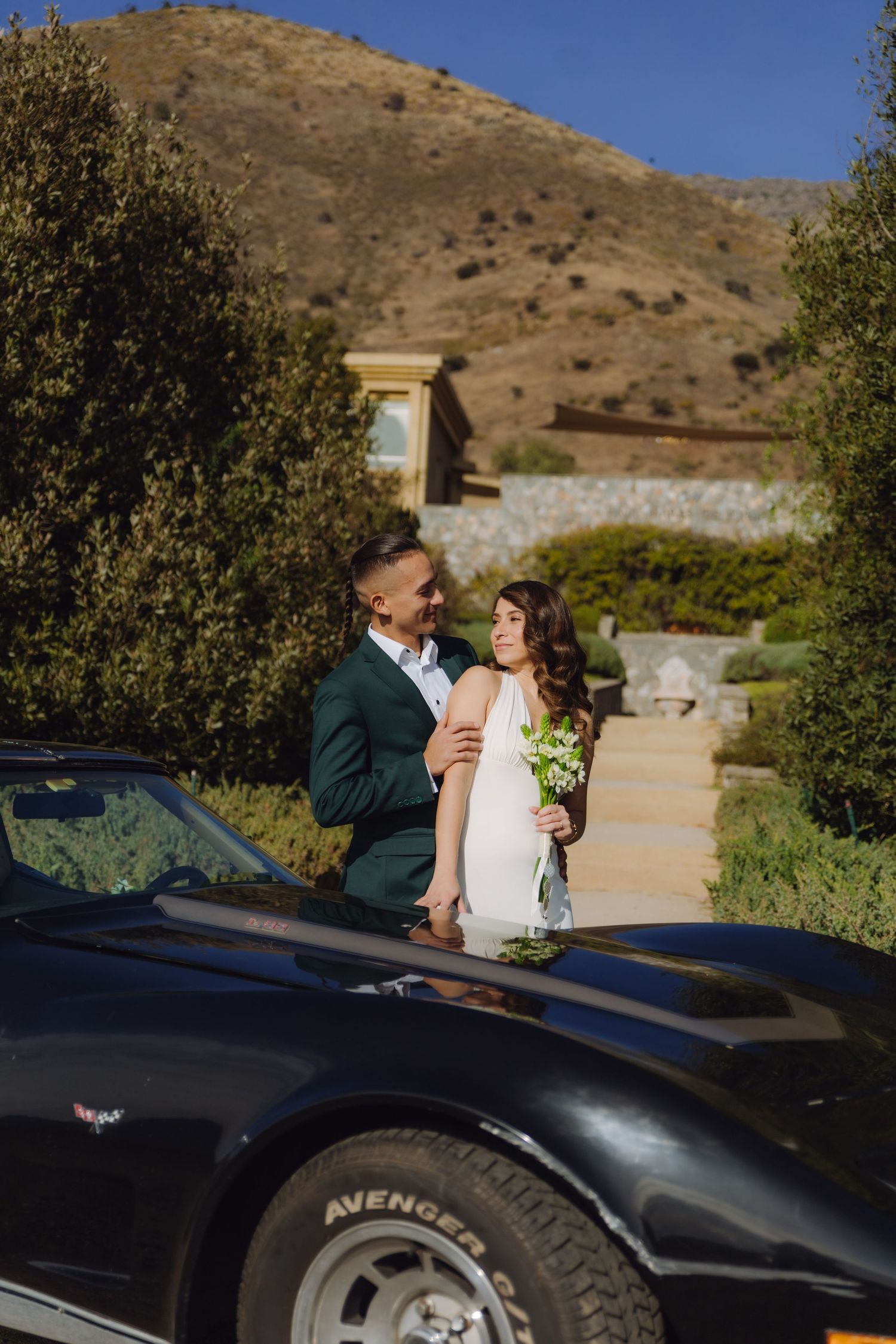 Smiling newlyweds embracing beside a black classic Corvette with lush greenery and hills.
