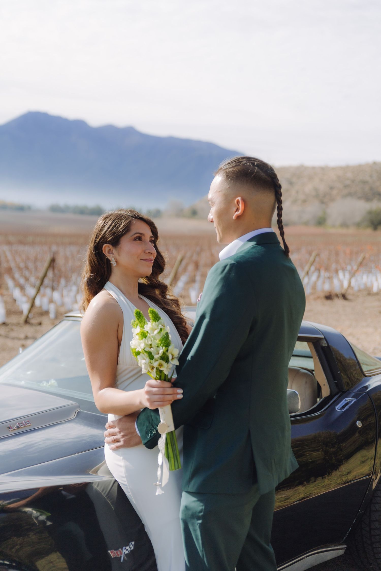 Bride holding white bouquet faces groom beside black Corvette with vineyard in background.