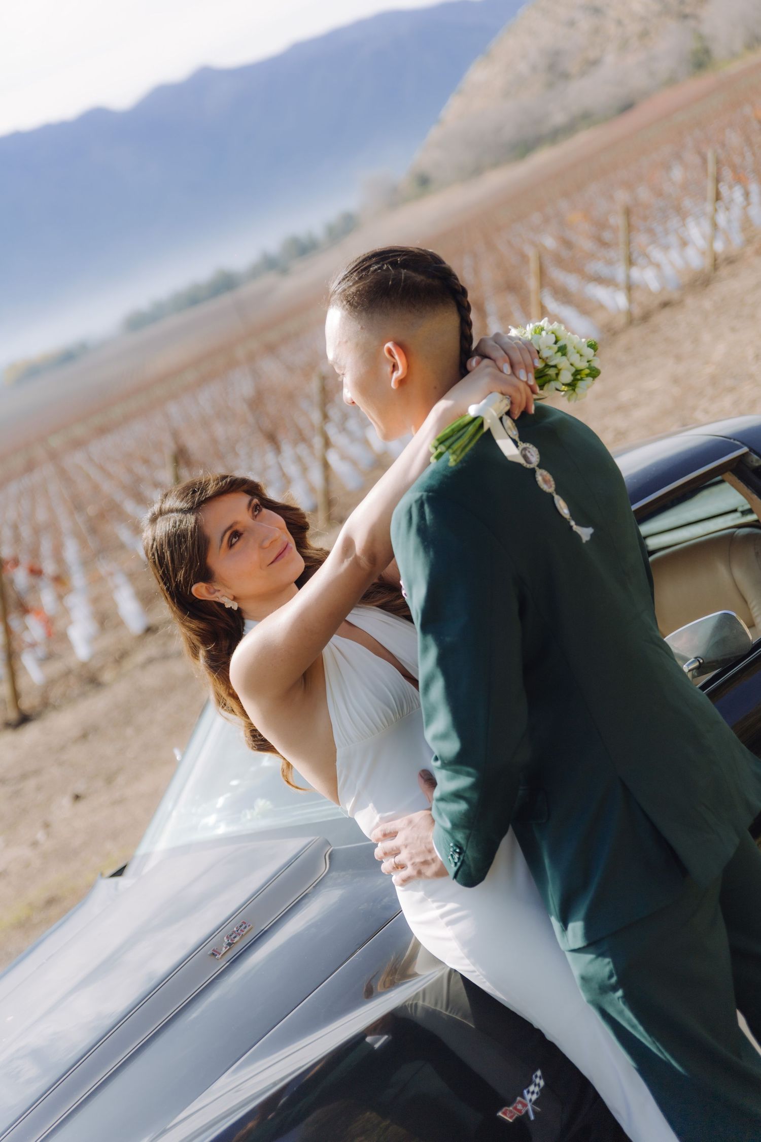 Bride embracing groom beside black Corvette with dormant winter vineyard in the background.