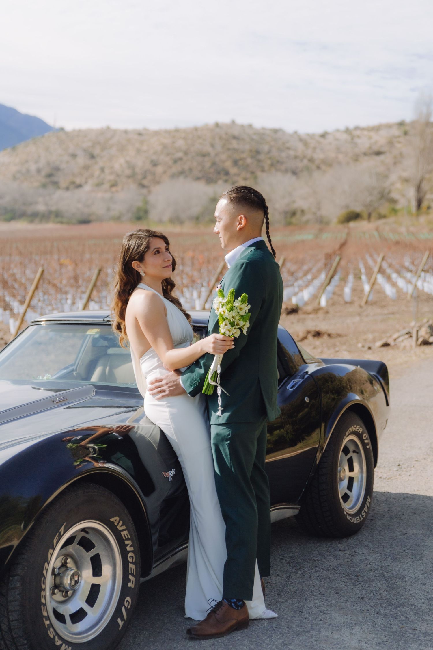 Newlyweds sharing an intimate moment leaning against a black Corvette near a winter vineyard.