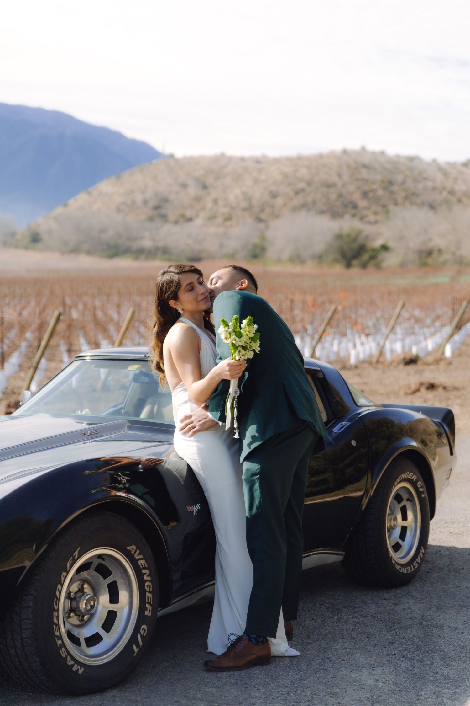Groom kissing bride beside a black vintage Corvette with a scenic vineyard backdrop.