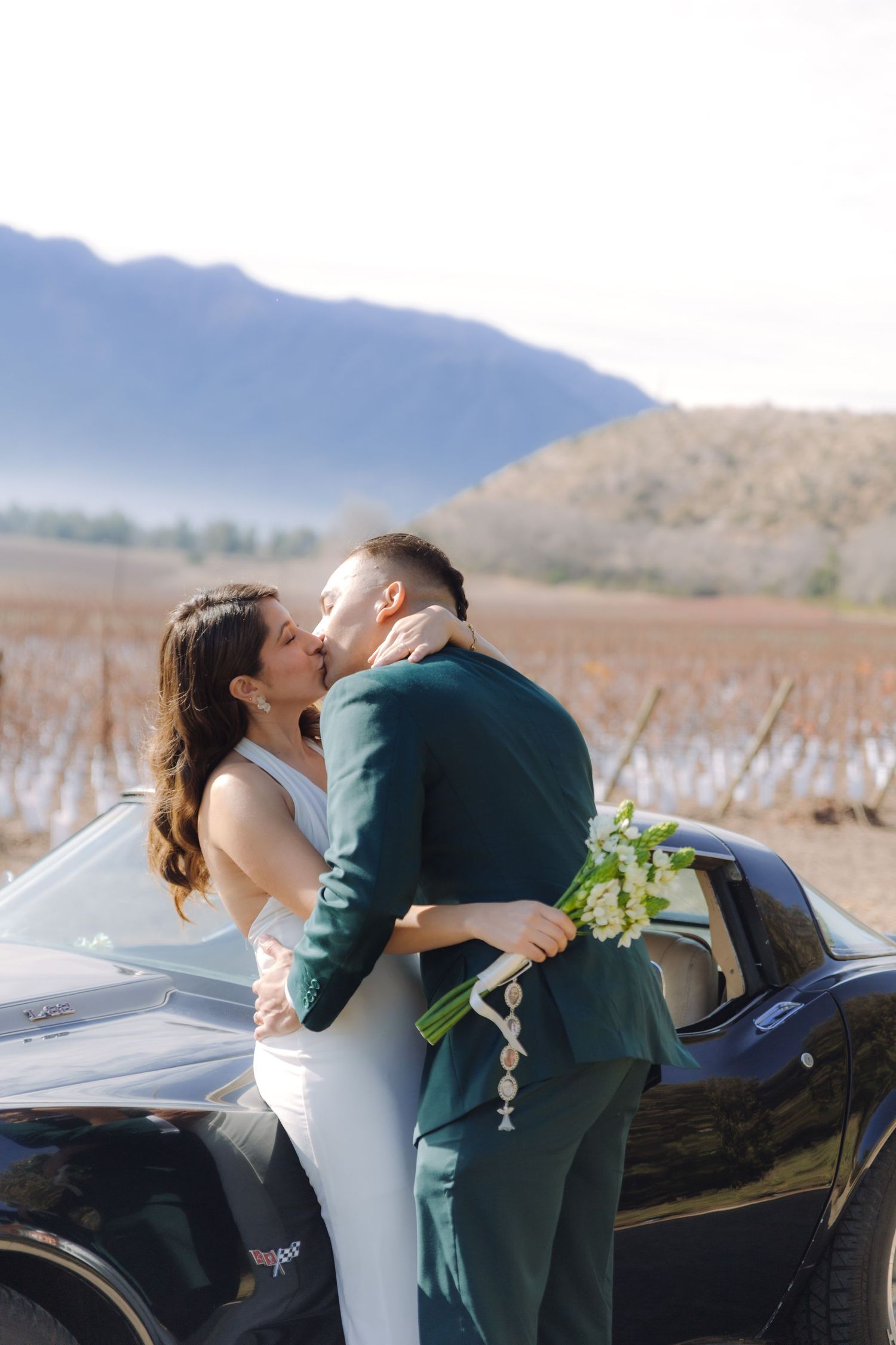 Bride and groom sharing a passionate kiss beside a black Corvette near a winter vineyard.