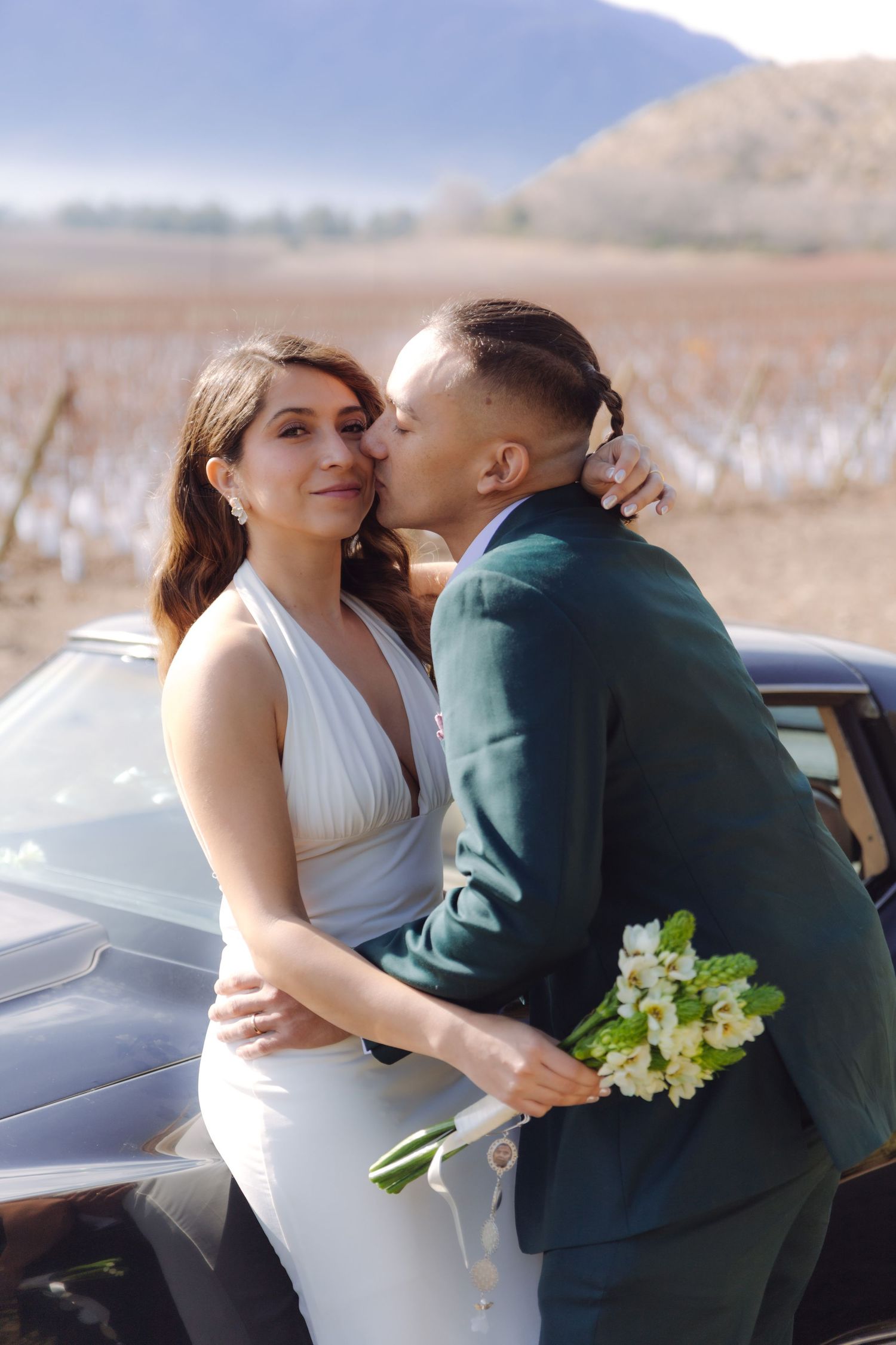 Groom in green suit kisses bride in white halter dress on cheek beside vintage car with mountain backdrop.