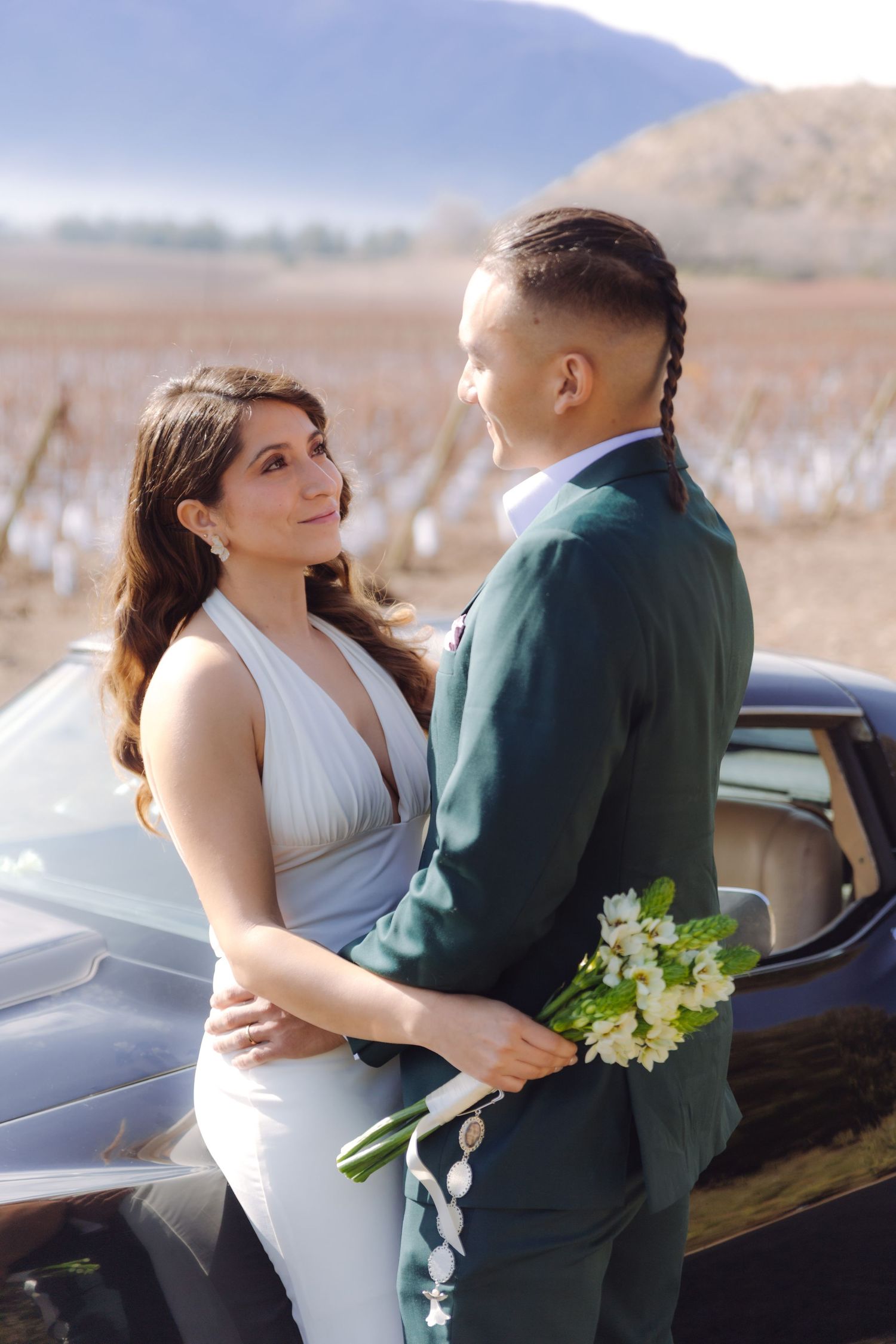 Wedding couple embracing by vintage black car, bride in white dress holds groom in green suit with bouquet.