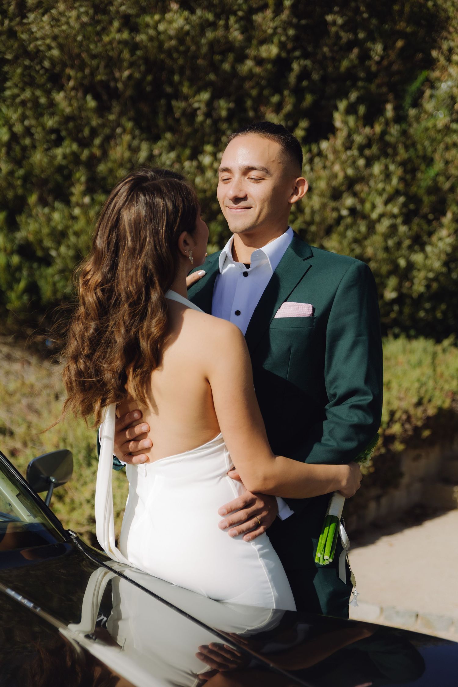 Smiling groom in green suit embraces bride in backless white wedding dress near a classic black car.