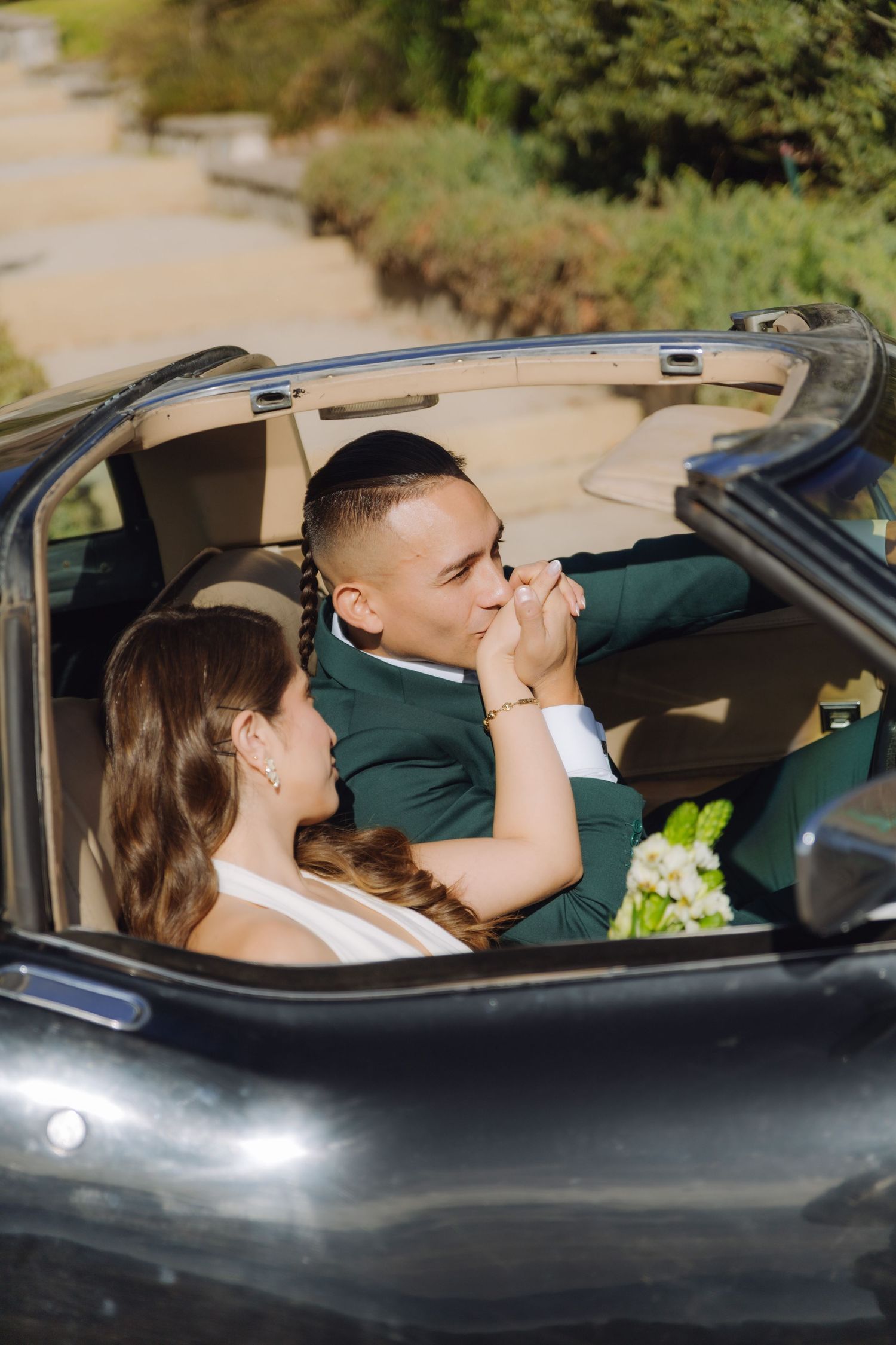 Groom in green suit kisses bride's hand in vintage convertible car during romantic wedding photo session.