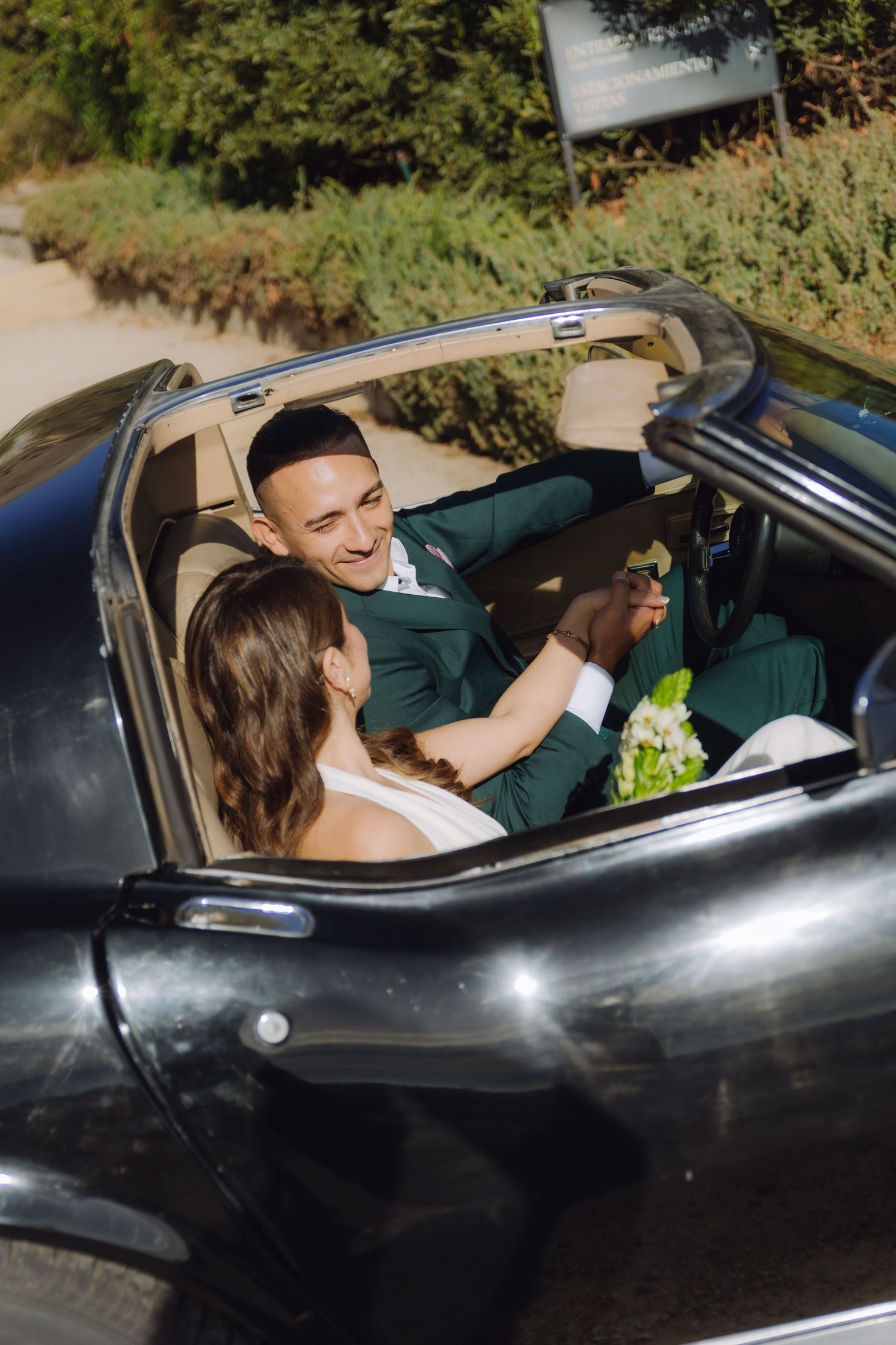 Smiling groom in green suit holds bride's hand inside a classic black convertible on their wedding day.