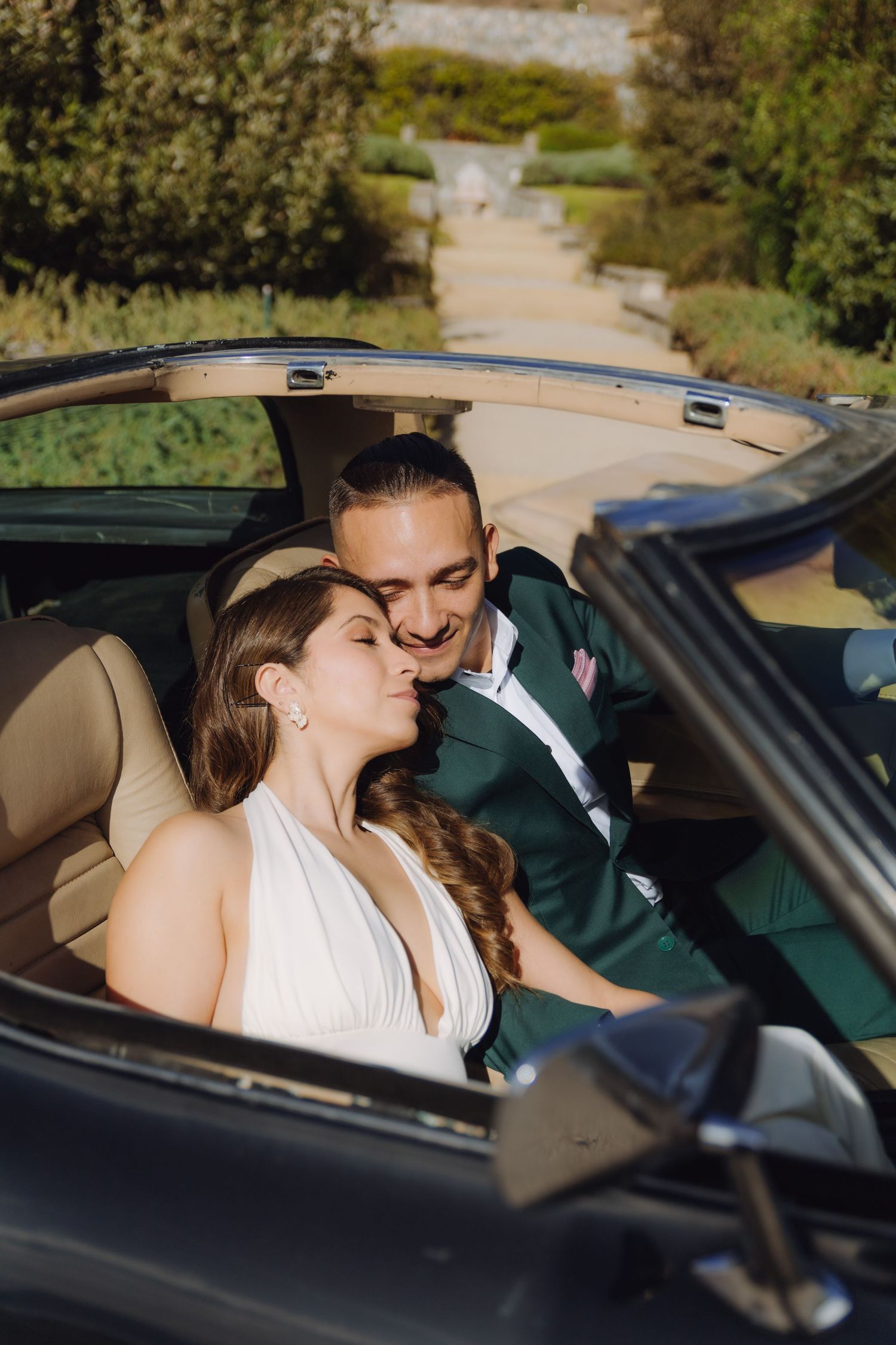 Bride and groom share a tender kiss while seated together in a vintage black convertible car.