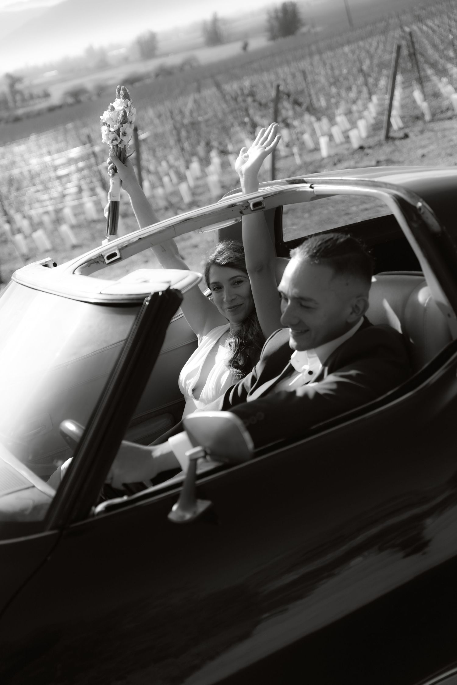 Black and white photo of newlyweds in vintage convertible, bride raising bouquet joyfully in vineyard setting.