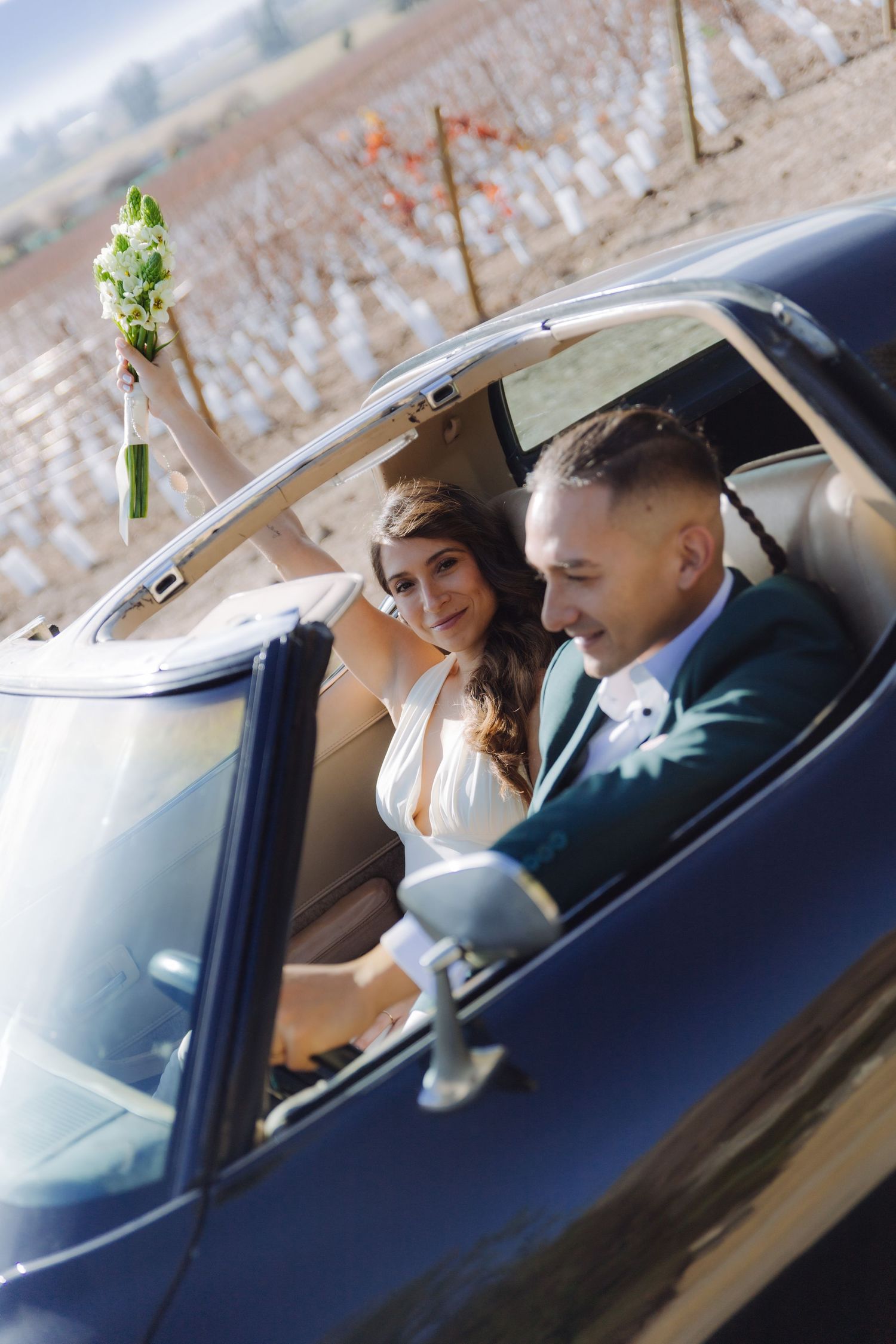 Bride raises floral bouquet while groom drives classic black convertible through scenic vineyard landscape.