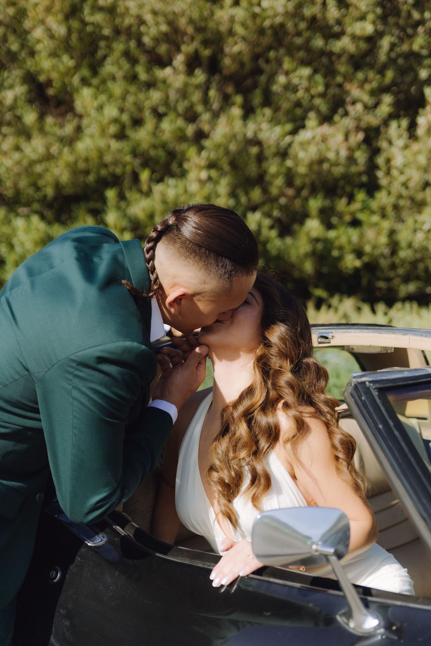 Bride and groom sharing a romantic kiss beside a vintage black convertible car outdoors.