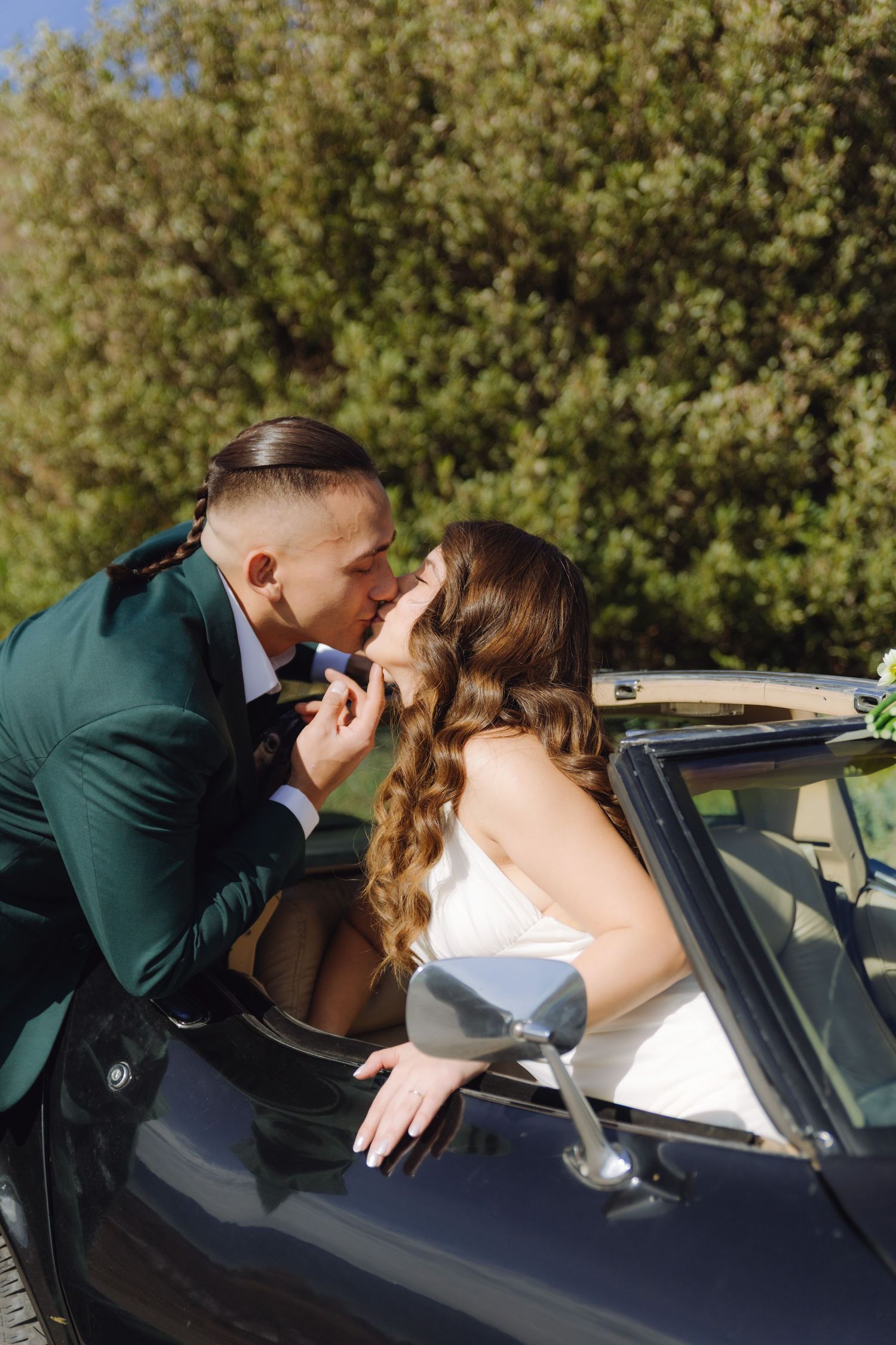 Wedding couple kissing near a classic black convertible car surrounded by lush green trees.