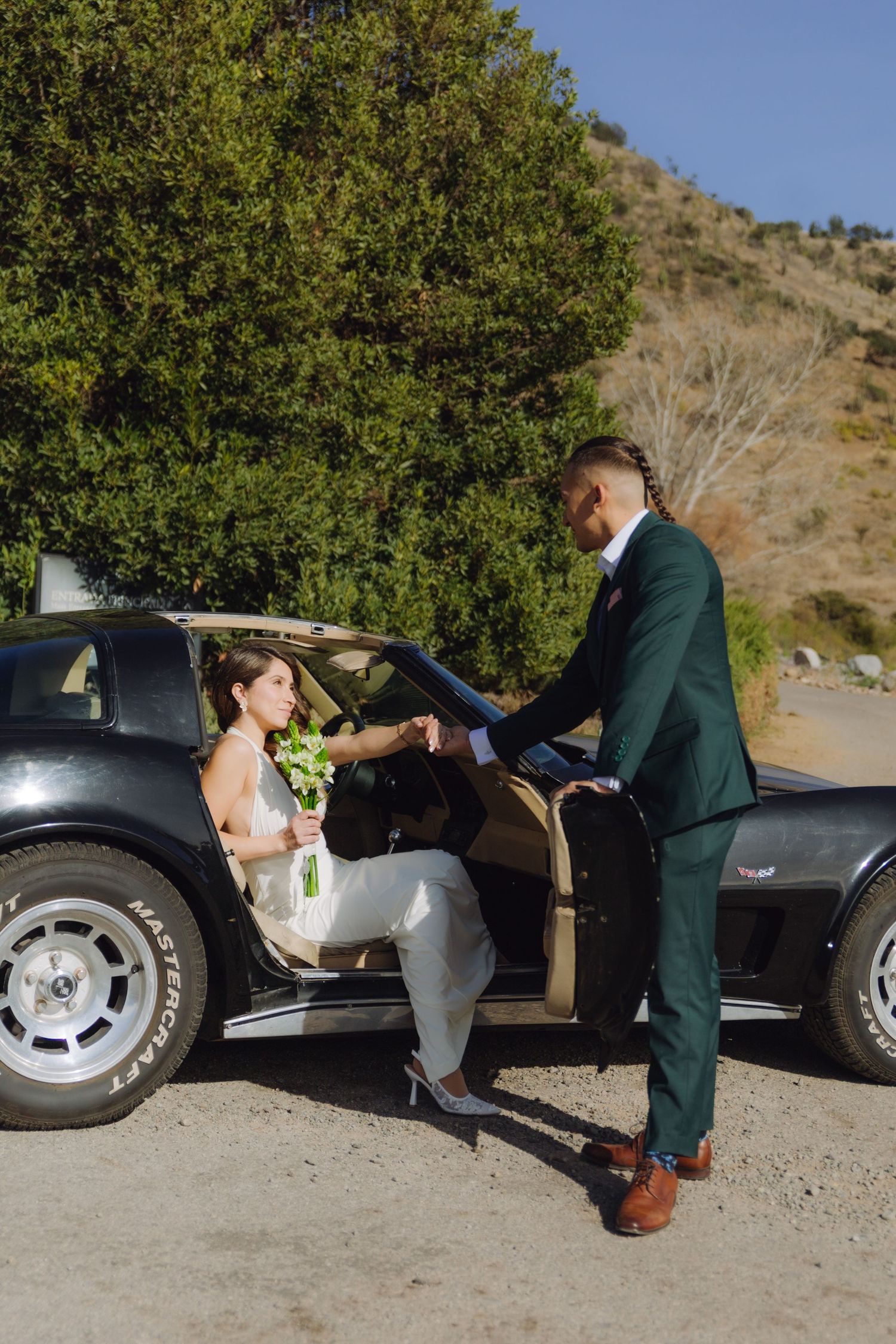 Groom in green suit helping bride out of a vintage black Corvette on a sunny day.