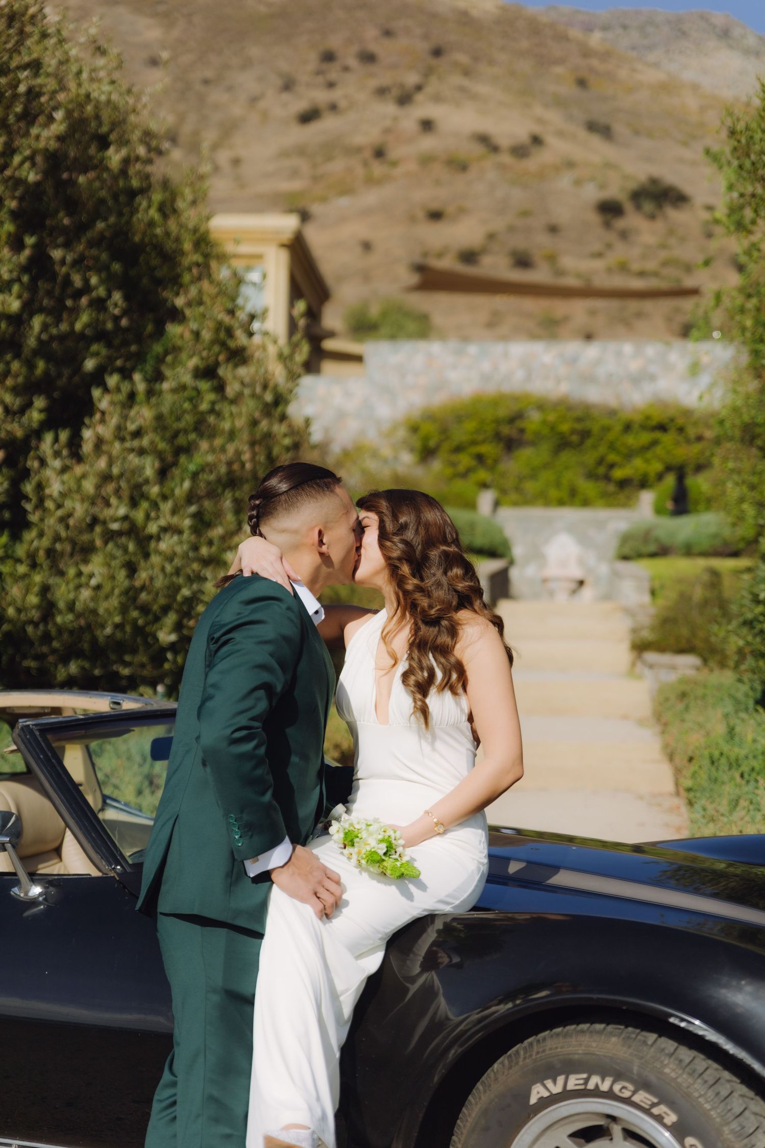 Bride and groom kissing beside a classic black convertible car with mountain backdrop.