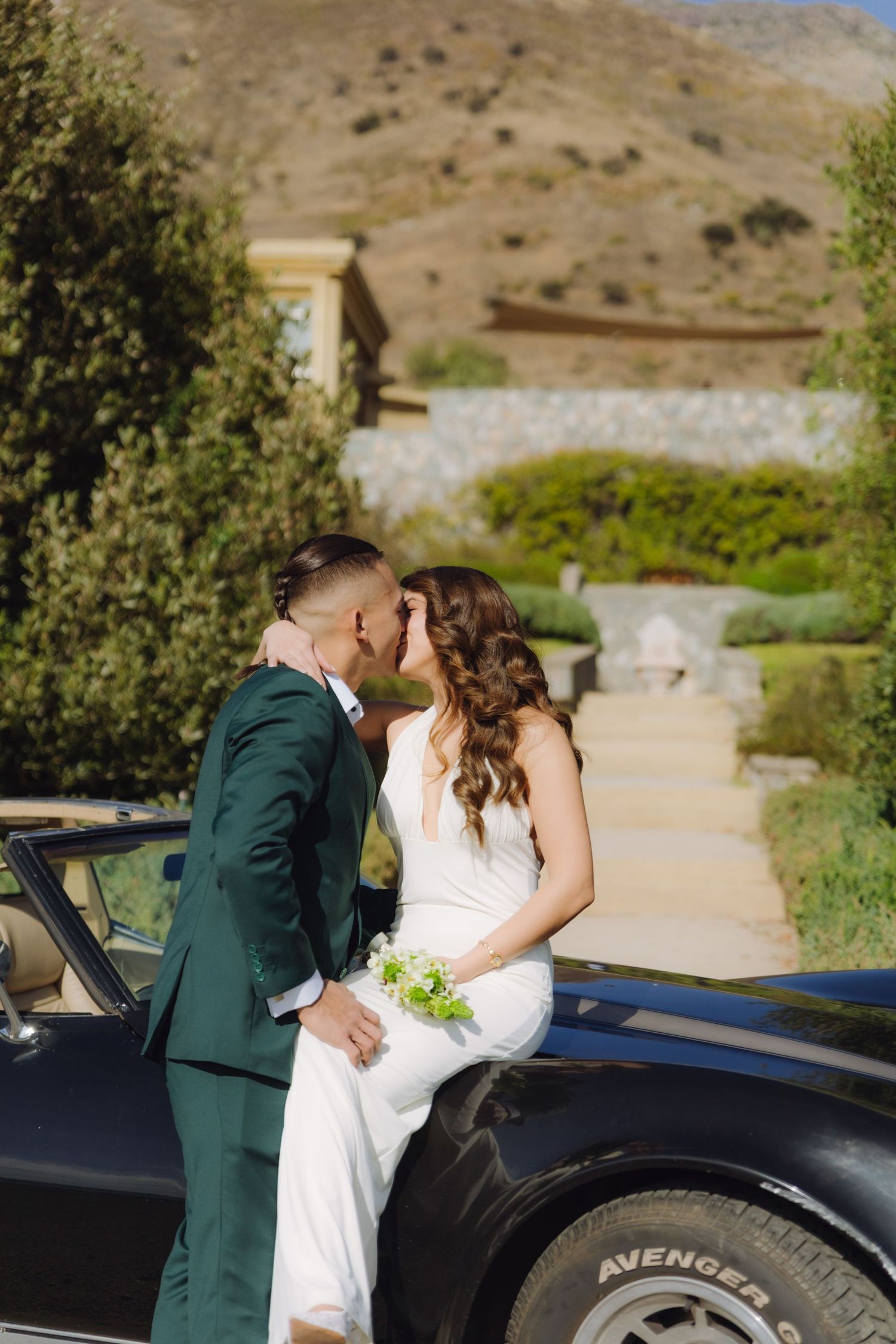Newlyweds sharing a kiss leaning against a vintage black convertible at an outdoor venue.