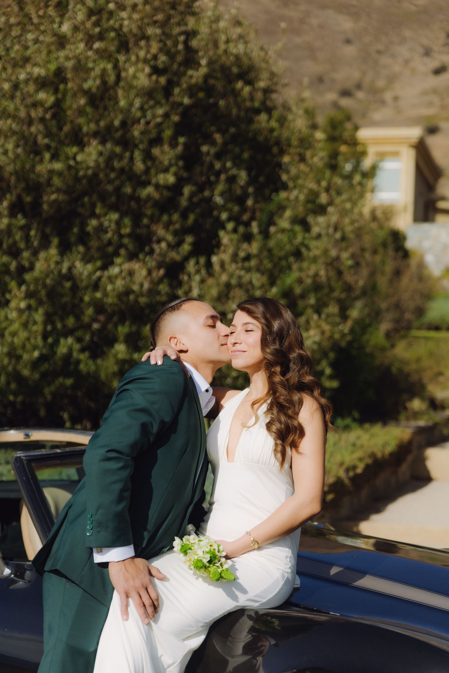 Groom in emerald suit kissing bride on cheek as she sits on a classic black car.