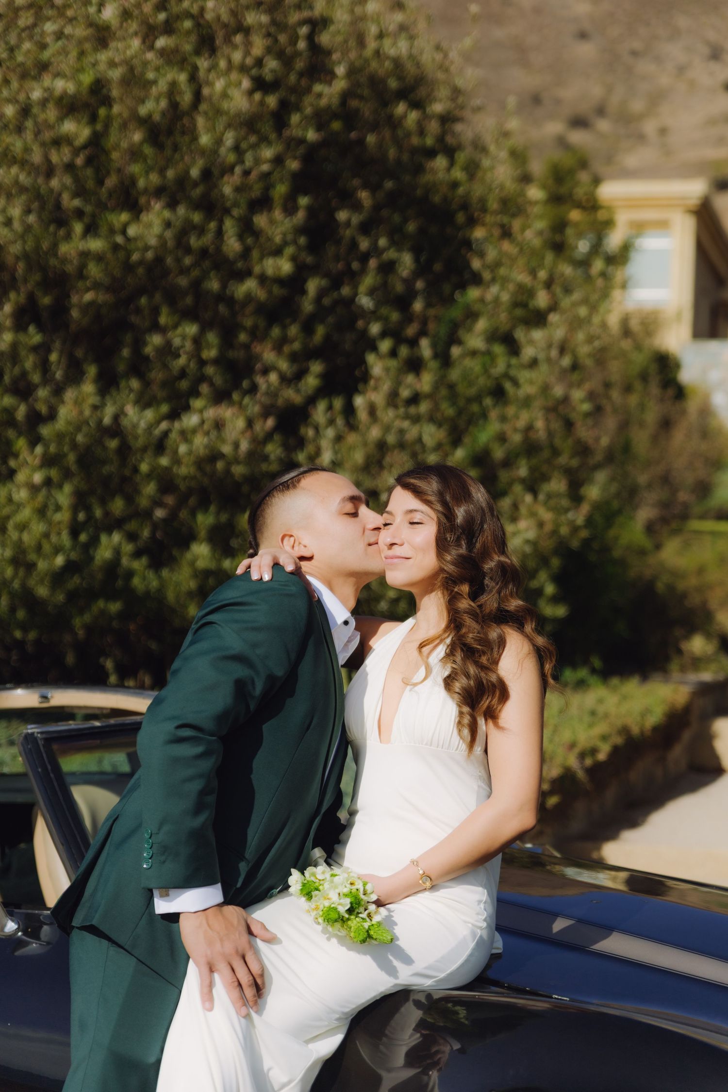 Bride holding green bouquet sits on vintage black car as groom kisses her cheek outdoors.