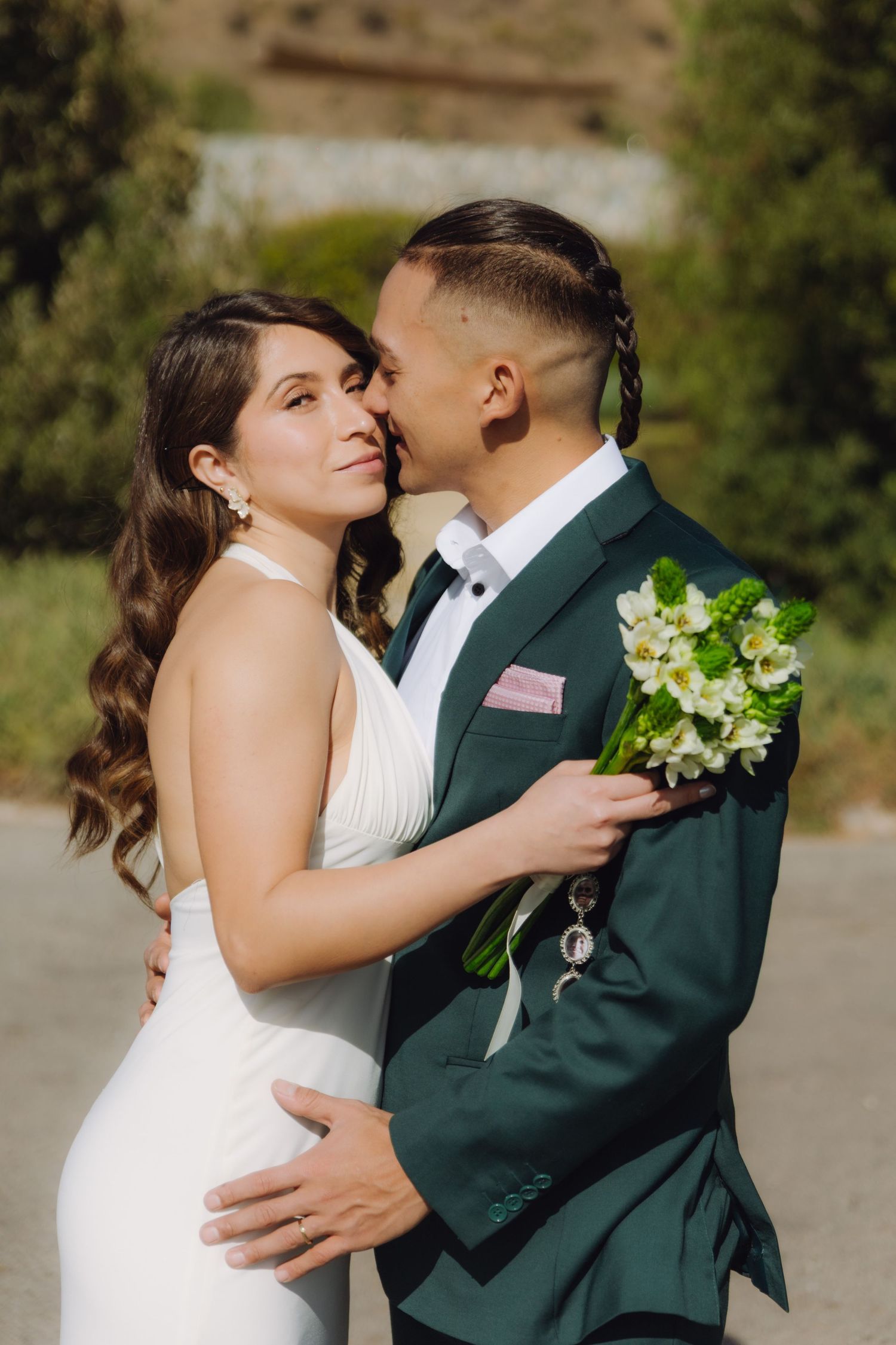 Bride and groom embracing closely outdoors, groom holding a white floral wedding bouquet.