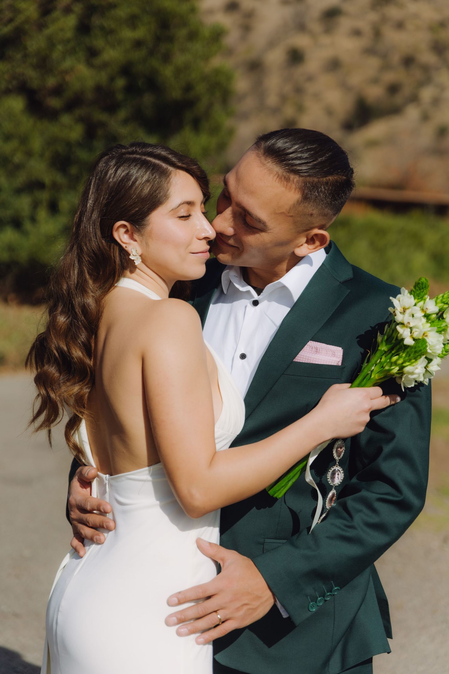 Bride and groom share a romantic embrace outdoors, she in a white halter gown, he in a dark green suit holding a bouquet.
