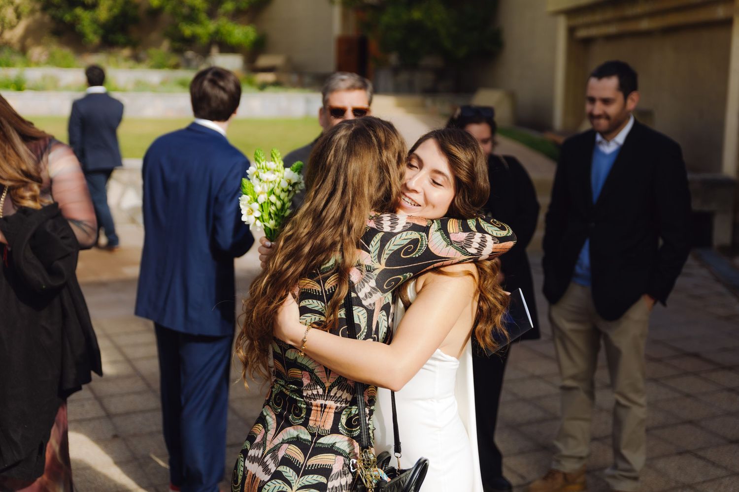 Wedding guests share warm hugs outside a venue, with a bride in white receiving an embrace from a woman in a floral dress.