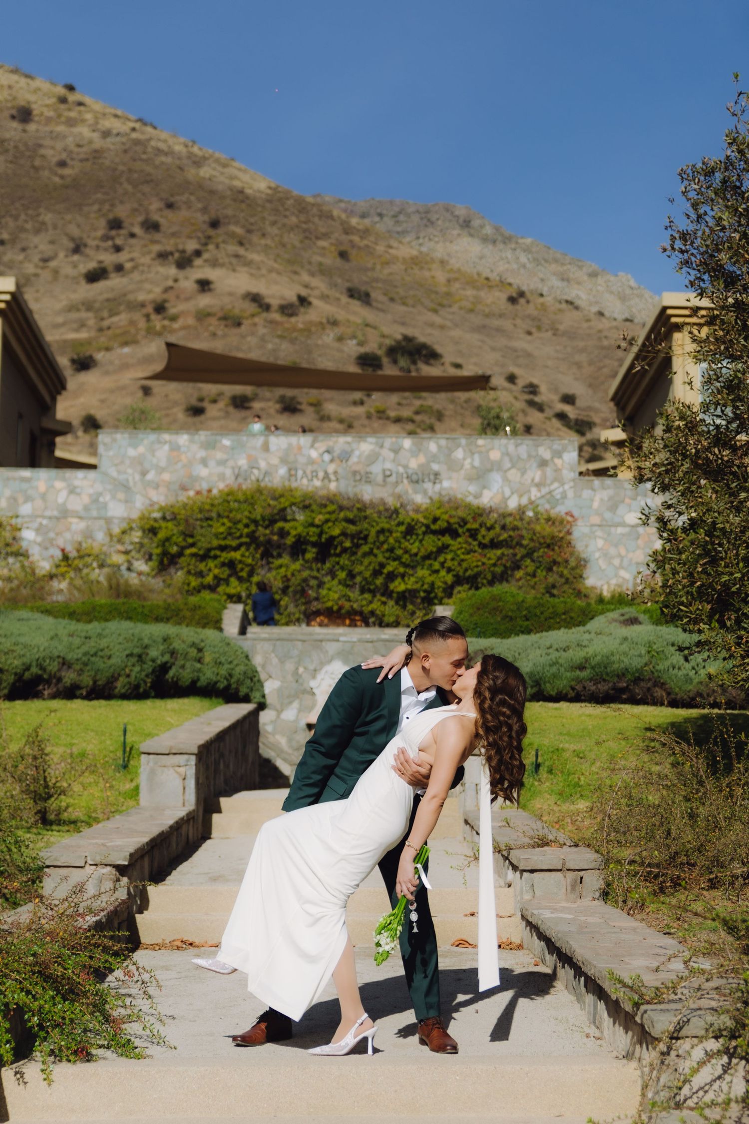 Bride and groom share a romantic dip kiss on garden steps with a stunning mountain backdrop on their wedding day.