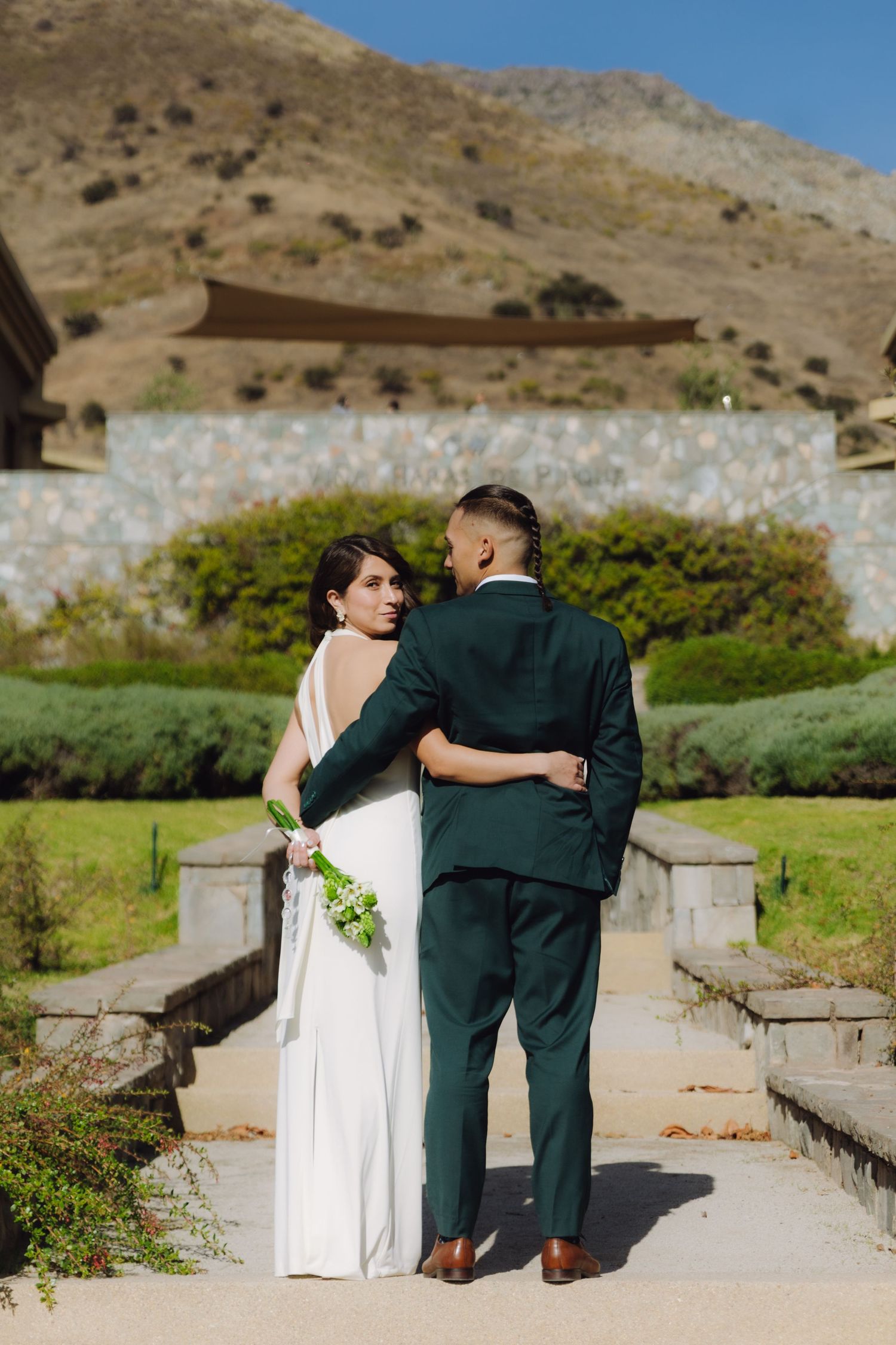 Bride in white backless gown and groom in green suit walk arm in arm toward a stone building with mountain views.