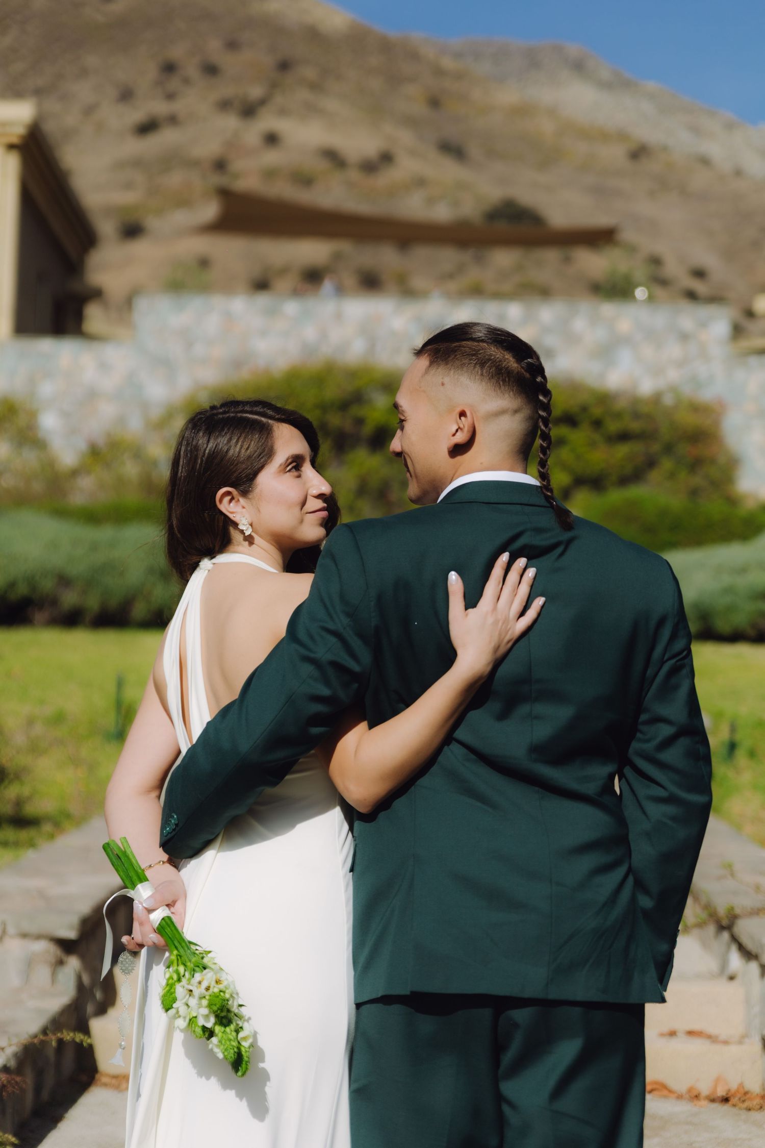 Bride in white halter dress and groom in dark green suit share a tender moment with a mountain backdrop.