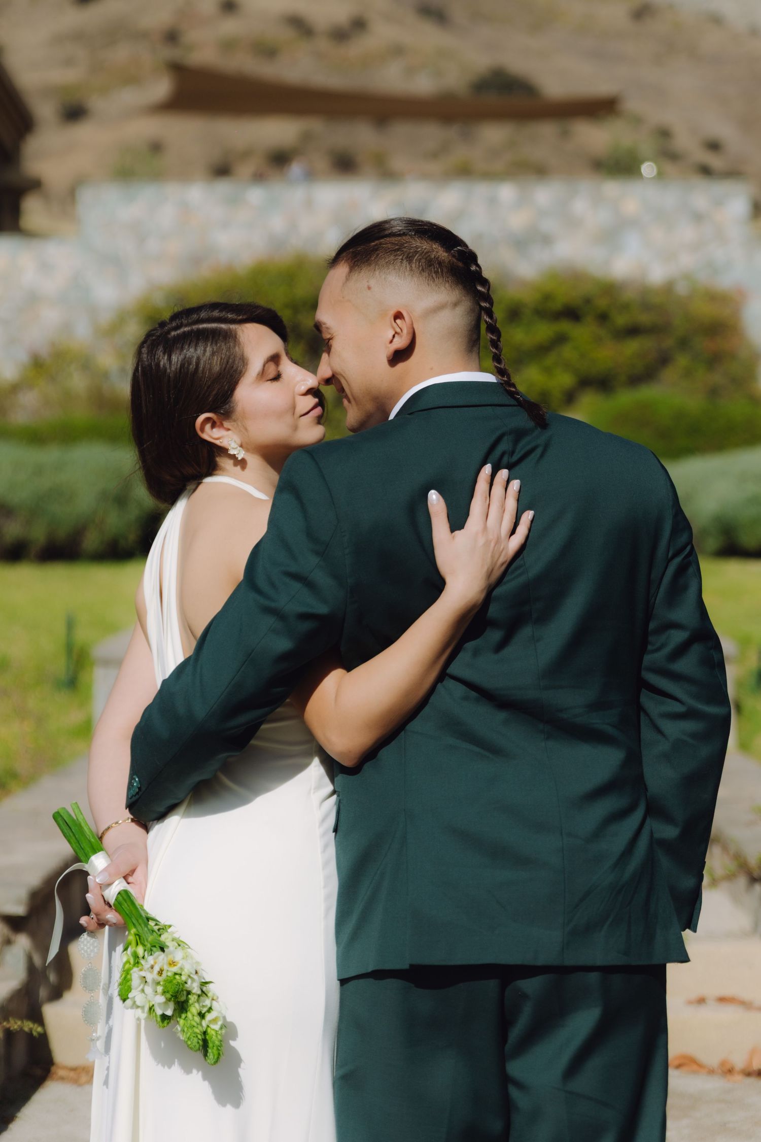 Bride and groom share an intimate nose-to-nose moment outdoors, surrounded by lush greenery and a mountain backdrop.