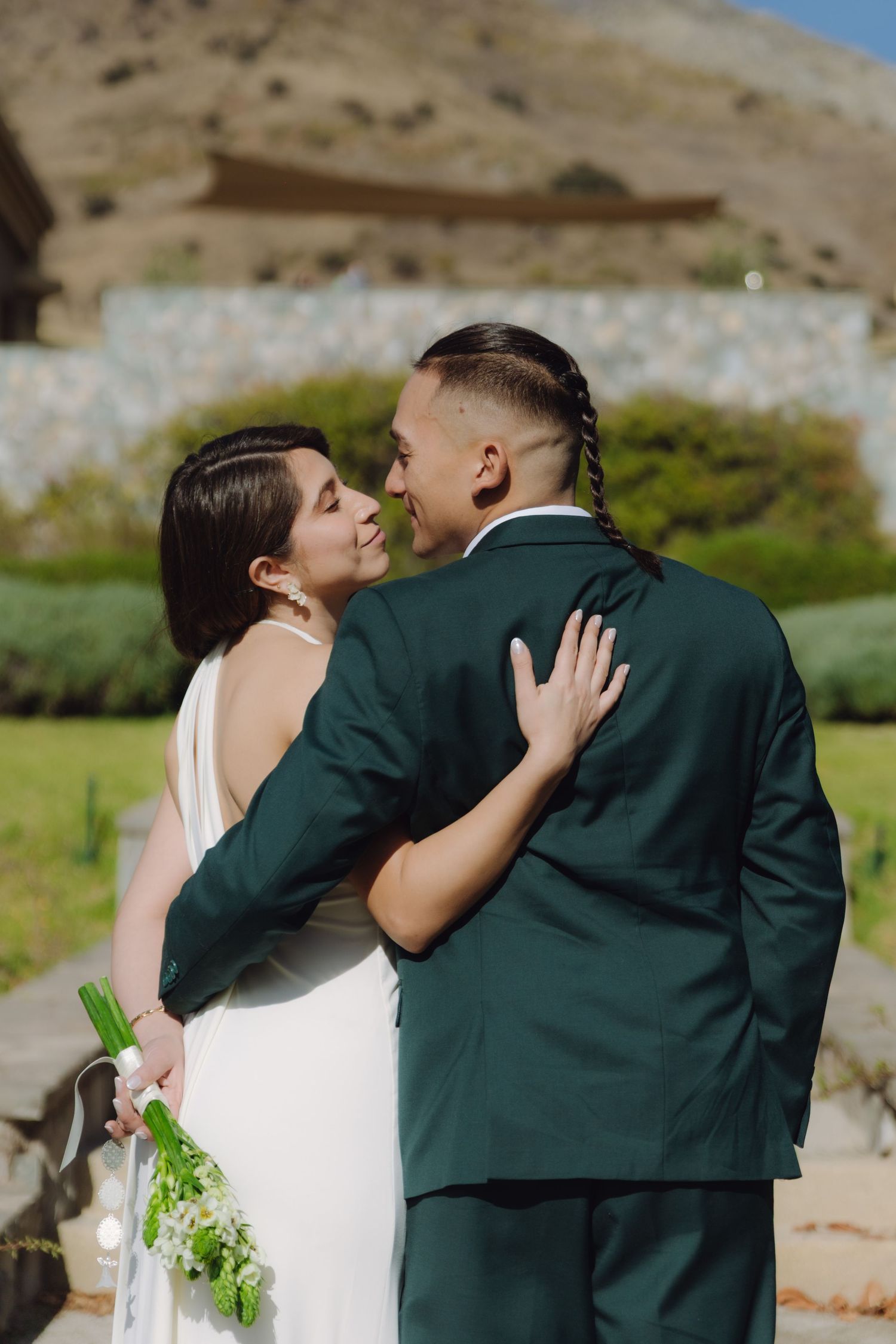 Bride and groom share a kiss outdoors, she in a white halter gown holding a bouquet, he in a dark green suit.